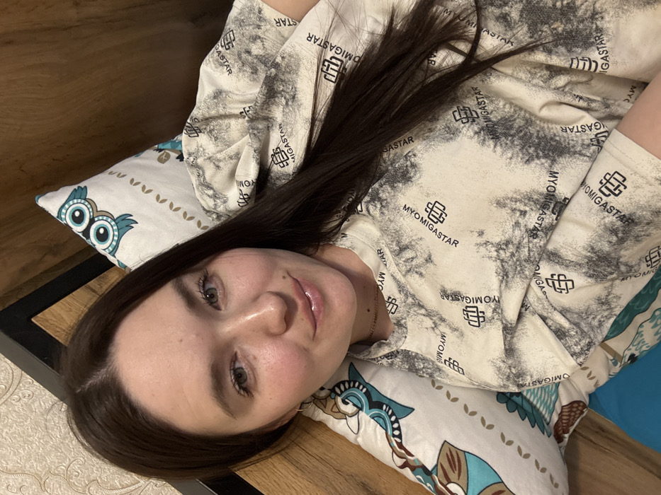 The scene is an interior location in Chaikovsky, Russia, likely a private residence. A young adult female is depicted lying down, her head resting on a white pillow adorned with a teal, brown, and off-white owl design. She has long dark brown hair, light-colored eyes, and a light complexion, with her lips appearing glossy and a subtle smile. She wears a light-colored, long-sleeved top featuring a grey mottled pattern interspersed with repeating "MYOMIGASTAR" text and a geometric logo. A small hoop earring is visible in her left ear, and a thin gold chain necklace is worn around her neck. She maintains direct eye contact with the camera. A wooden headboard or bed frame is visible behind her, with a dark, possibly metallic, frame element at the top left. Light-colored patterned wallpaper is partially visible in the upper left background.