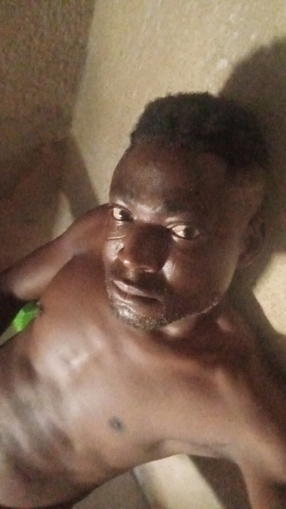 A medium shot of a shirtless, dark-skinned man with short, dark hair and a trimmed beard. He is looking directly at the camera with a slightly upward gaze. His left arm is raised with his hand near his face. The background is a blurred beige color, possibly a wall. It's unclear from the image if the location is Sabon Tasha, Nigeria.