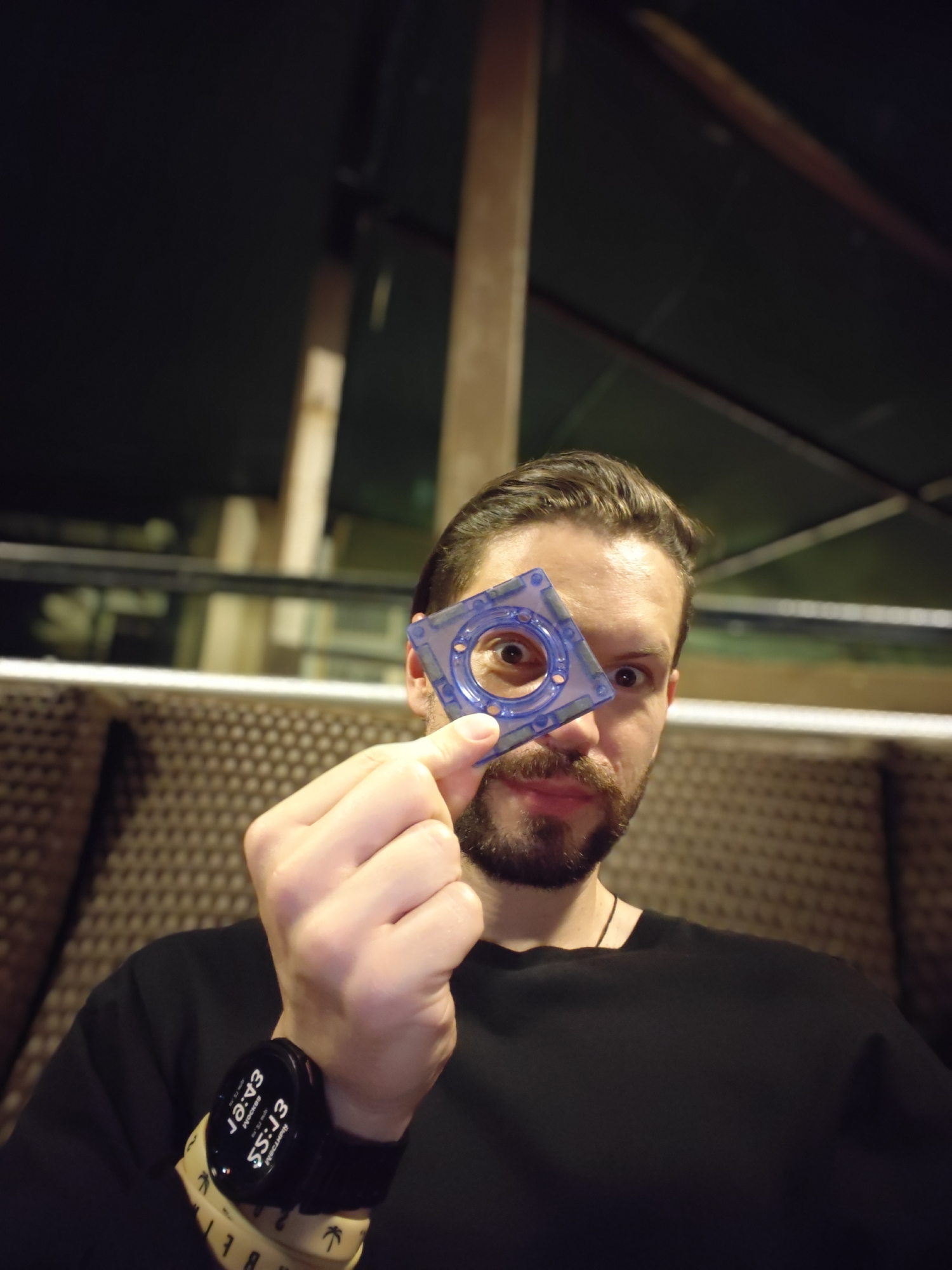 The image features a man with a beard and dark hair, wearing a black t-shirt, captured in an indoor setting in Mumbai, India. He is looking directly at the camera with a wide-eyed, playful expression, holding a transparent blue, square-shaped object with a circular cutout in the center over his right eye. The object appears to be a piece of a magnetic building toy or puzzle.
His left wrist is visible, adorned with a black smartwatch displaying "12:43" and "55:13". Below the smartwatch, he wears two wristbands: one yellowish-beige with the partially visible text "BUREAU", an upward arrow, and "FT:SS 55:13", and another black wristband featuring palm tree designs.
The background is dimly lit, suggesting an evening or night ambiance. On the right side behind the man, a textured, possibly padded or woven, brown wall or partition is visible. To the left and above, dark areas contrast with what appear to be wooden beams or structural elements of the building. The overall scene conveys a casual and relaxed atmosphere.