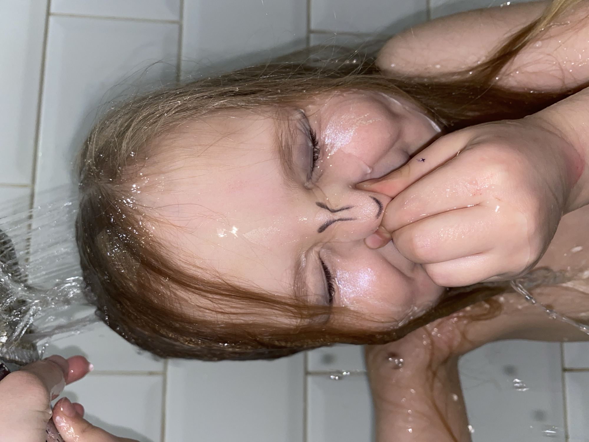 A young child with light brown hair is in a bathtub under a showerhead. The child's eyes are closed, and water is streaming over their face and hair. There is a drawing of a cat's nose and whiskers on the child's face. The child is pinching their nose with one hand. The background consists of white tiles. There is no indication that the image was taken in the city of Votkinsk, Russia.