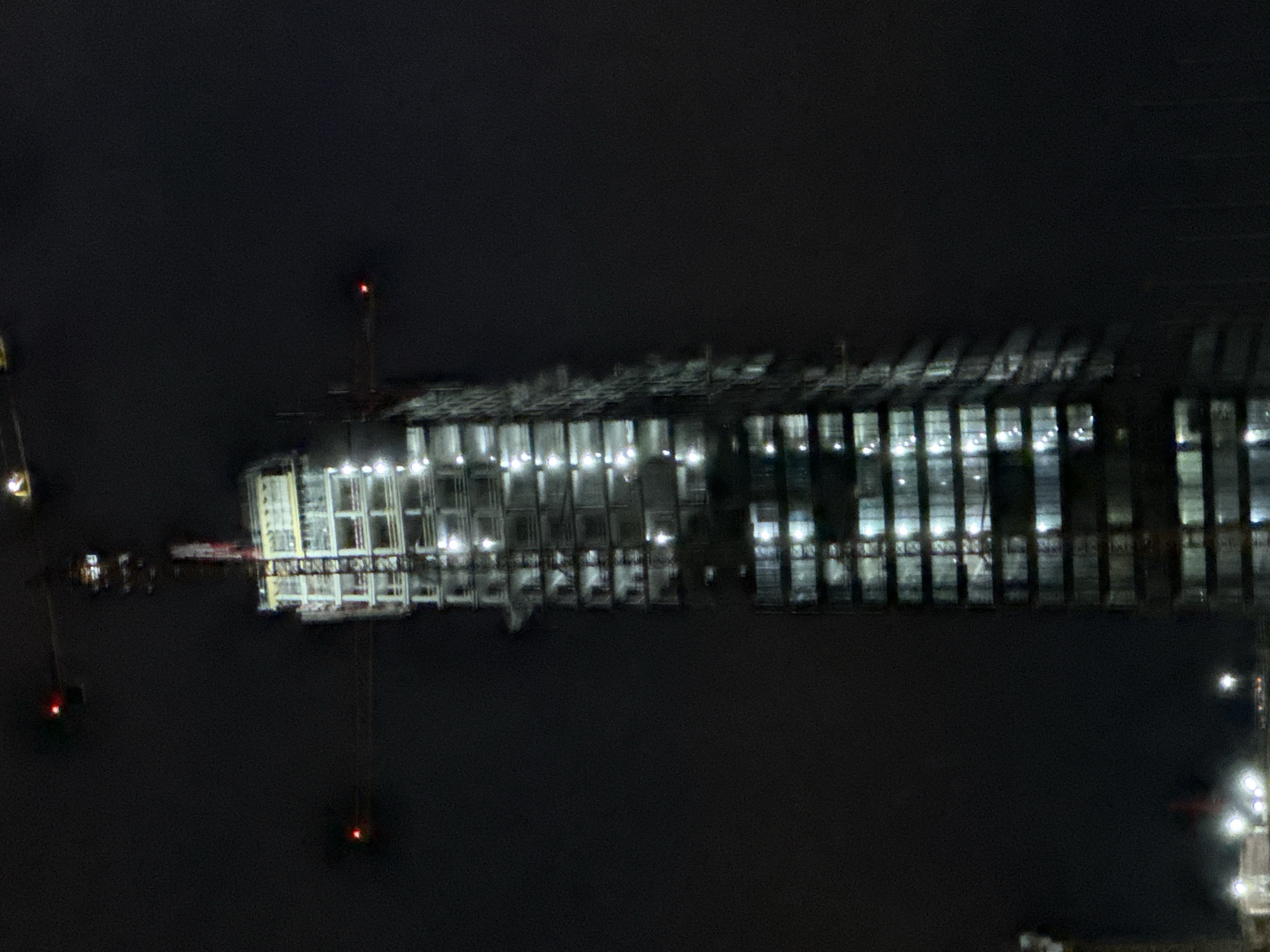 An eye-level, full shot captures a towering skyscraper under construction at night. The structure is illuminated with numerous bright lights, highlighting its skeletal framework. Cranes, characteristic of a construction site, are visible above and around the building. In the distance, more lights suggest the presence of other buildings in a city. The sky is dark and hazy, obscuring any celestial details and creating a moody atmosphere.
