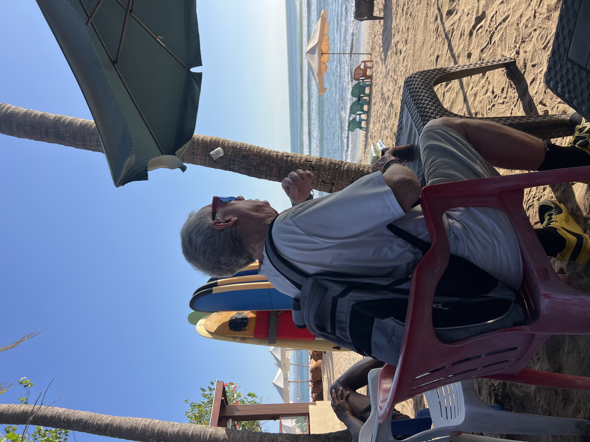 In Kuta, Indonesia, a Caucasian male with gray hair and red-framed sunglasses sits at a black woven table on a sandy beach. He wears a white t-shirt, light-colored shorts, black socks, and yellow athletic shoes. A gray and black backpack is on the back of the red plastic chair he occupies. His left arm rests on the table, holding a dark cup. Behind him, a palm tree trunk stands to the left, with a large green umbrella providing shade above. Several surfboards are propped against a building with a wooden facade to the left of the frame, where white beach umbrellas and brown beanbag chairs are visible. In the background, the ocean with waves meets a clear blue sky, and other beach chairs and umbrellas are scattered across the sand.