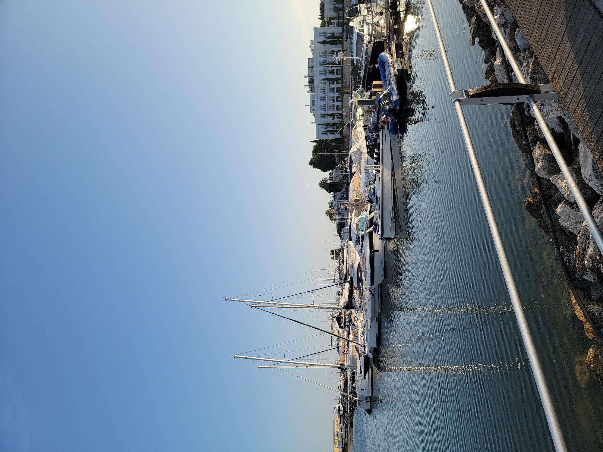 This is a vertically oriented photograph taken at a marina in Quarteira, Portugal. The scene depicts a row of moored sailboats and yachts docked along a wooden pier. The water in the marina is calm, with gentle ripples reflecting the bright, clear sky.
In the background, a series of white buildings with multiple floors are visible, suggesting a coastal town or resort. Some of these buildings are partially obscured by lush green trees. The sky is a clear, pale blue, indicative of a sunny day. The lighting suggests it is either early morning or late afternoon, given the long shadows cast by the boats and the warm glow on the water.
The boats are of varying sizes, with masts of some sailboats prominently extending upwards. Some of the yachts have their covers on, indicating they are either not in use or being protected from the elements. A few smaller inflatable boats are also docked alongside the larger vessels.
A wooden walkway runs alongside the marina, bordered by natural stone rocks. Metal railings are installed along the walkway, with a few stanchions and ropes visible. There are no people actively engaged in activities on the boats or the pier, giving the scene a tranquil and peaceful atmosphere. The overall impression is one of a serene and picturesque marina setting.