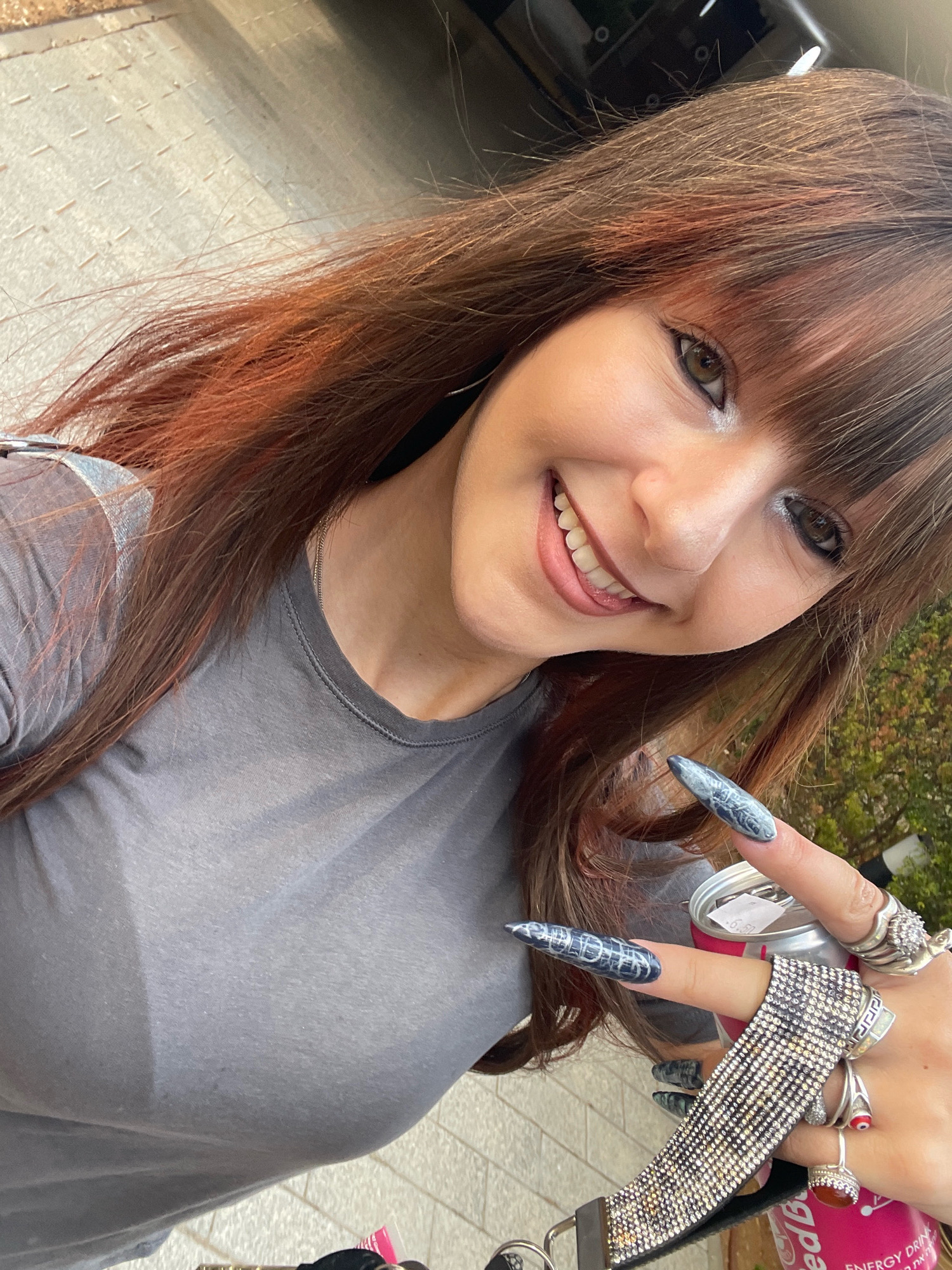 The image shows a young woman with long, brown hair with reddish highlights, bangs, and dark eye makeup. She is smiling and holding a can with her right hand, which is adorned with several rings and long, dark, patterned fingernails. She is wearing a grey t-shirt and a silver necklace. The background appears to be an outdoor setting with a paved surface and some greenery. The image is cropped tightly around the woman's face and hand. Based on the presence of Hebrew text on the can which reads "Energy Drink" and some other text that appears to be in Hebrew, as well as the general appearance of the paved surface, it is plausible this image was taken in Raanana, Israel.