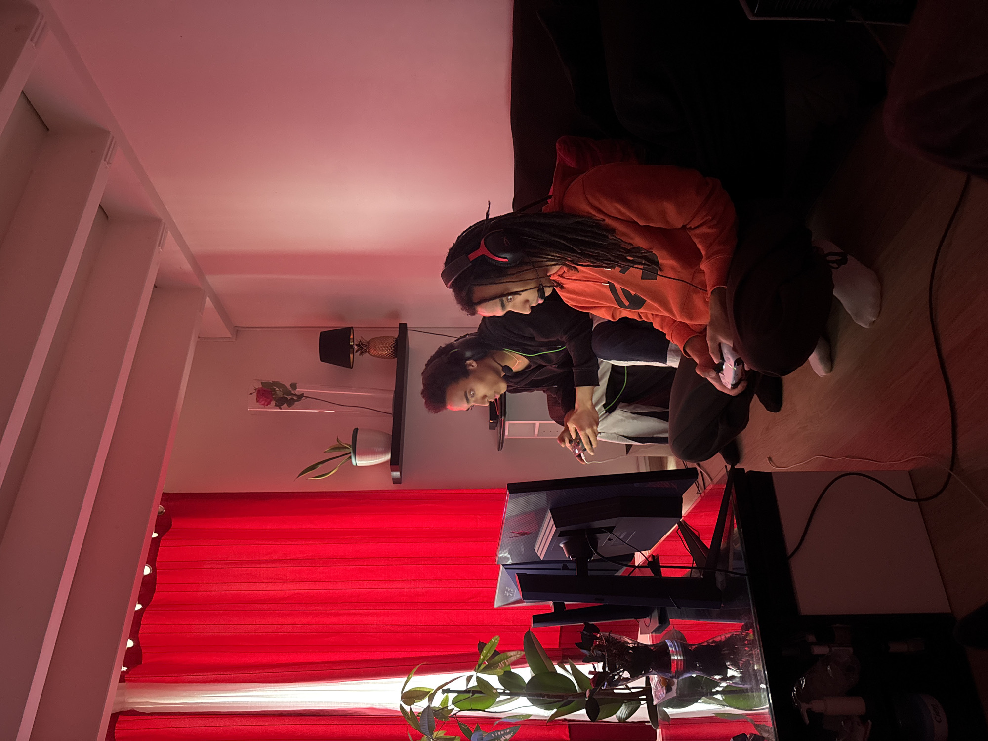 Two young men are playing video games in an apartment. The apartment has red curtains and a plant on a glass table. One of the men has dreadlocks and is wearing a red hoodie. The other man is wearing a black jacket with green trim. The location is not identifiable as Paris, France based on the visual information provided.
