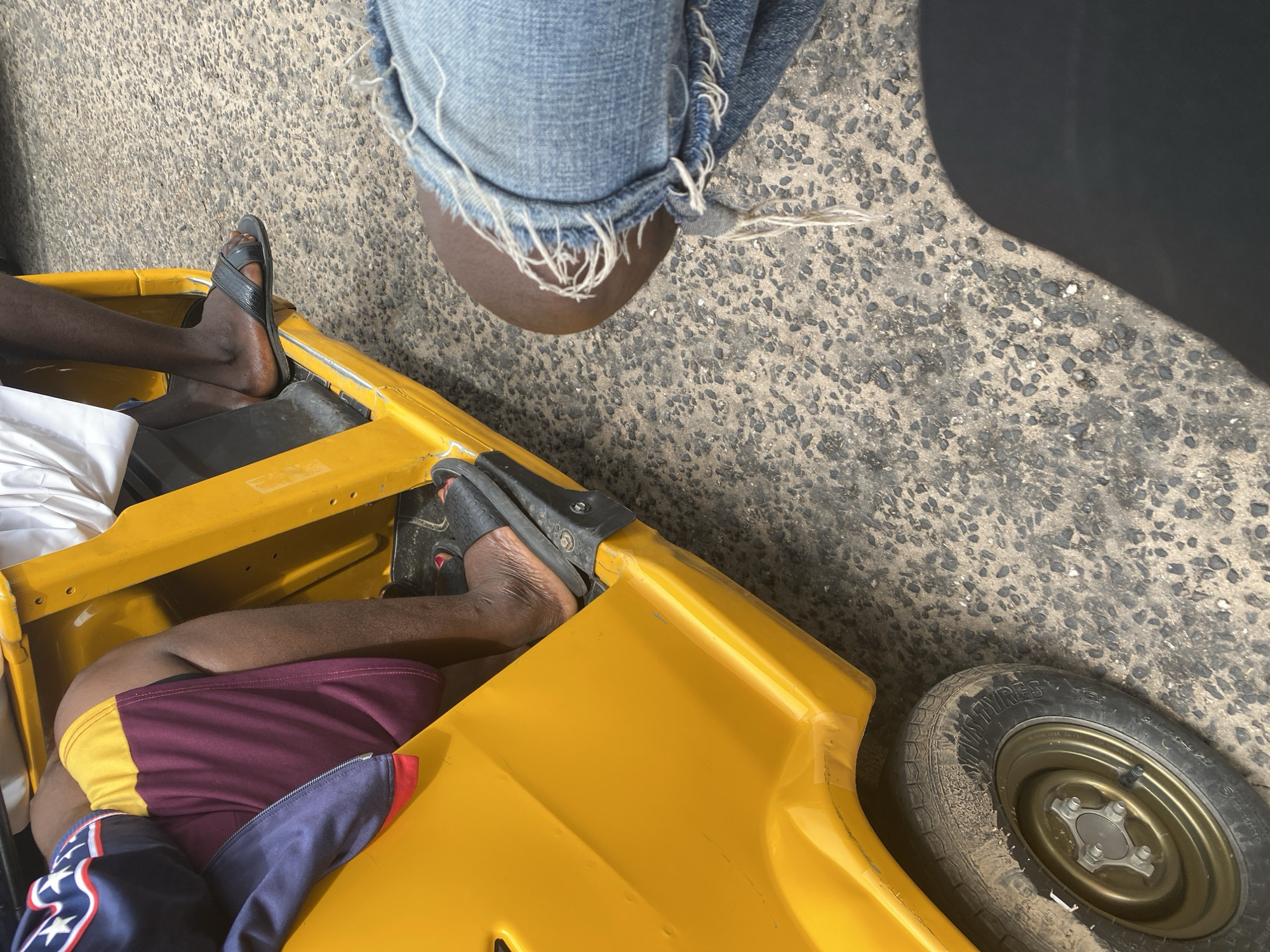 The image captures a close-up, overhead view of two individuals, their legs and feet visible, seated within a bright yellow vehicle, possibly a tuk-tuk or a similar open-air transport. The ground is a rough, grey asphalt surface, suggesting an outdoor setting. One person is wearing distressed denim jeans with frayed hems, and their feet are partially obscured by the fabric. The other individual is wearing shorts in a burgundy and yellow color combination, along with a dark blue garment with white stars and red trim draped over their legs.
One person's feet are adorned with black sandals. The other person's feet are bare, with painted red toenails visible. The yellow vehicle appears to be utilitarian, with visible edges and a portion of a spare tire on the right side of the frame. The tire has "TIRES" visible on its sidewall. A dark shadow falls across the right side of the image, indicating it's likely daytime, possibly under direct sunlight. The scene suggests a moment of rest or travel in a possibly urban or semi-urban environment in Jalingo, Nigeria. There is no discernible text beyond that on the tire.