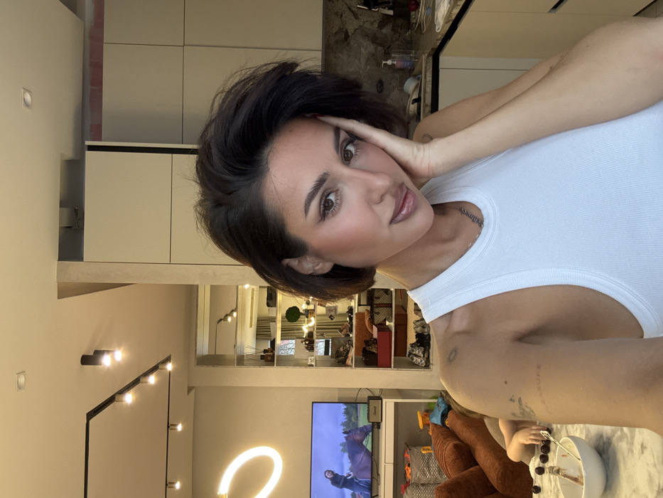 This image depicts an indoor scene within a modern apartment in Khimki, Russia. A female subject with short, dark hair, styled in a bob, and wearing a white ribbed tank top, occupies the foreground. She has visible makeup, and her left hand is resting against her cheek. A silver necklace and a faded tattoo, partially showing the text "PLEASURE", are visible on her left upper arm.
The background comprises a combined living and kitchen area. To the left, a flat-screen television displays an equestrian scene. Below it, a white console table holds a Marshall speaker and a gray basket. A neon O-shaped light illuminates the wall. A brown upholstered sofa is partially visible, with a partially obscured individual lying on or behind it. A white marble-patterned table in this area holds a bowl of cherries with a spoon, and additional cherries are scattered on the table surface.
To the right, a kitchen section features light-colored flat-panel cabinets, a marble backsplash, and a counter with various bottles. Beyond this, a mirrored shelving unit displays shoes, handbags (including a Louis Vuitton item), and decorative elements like a small green plant. Track lighting is installed on the ceiling.