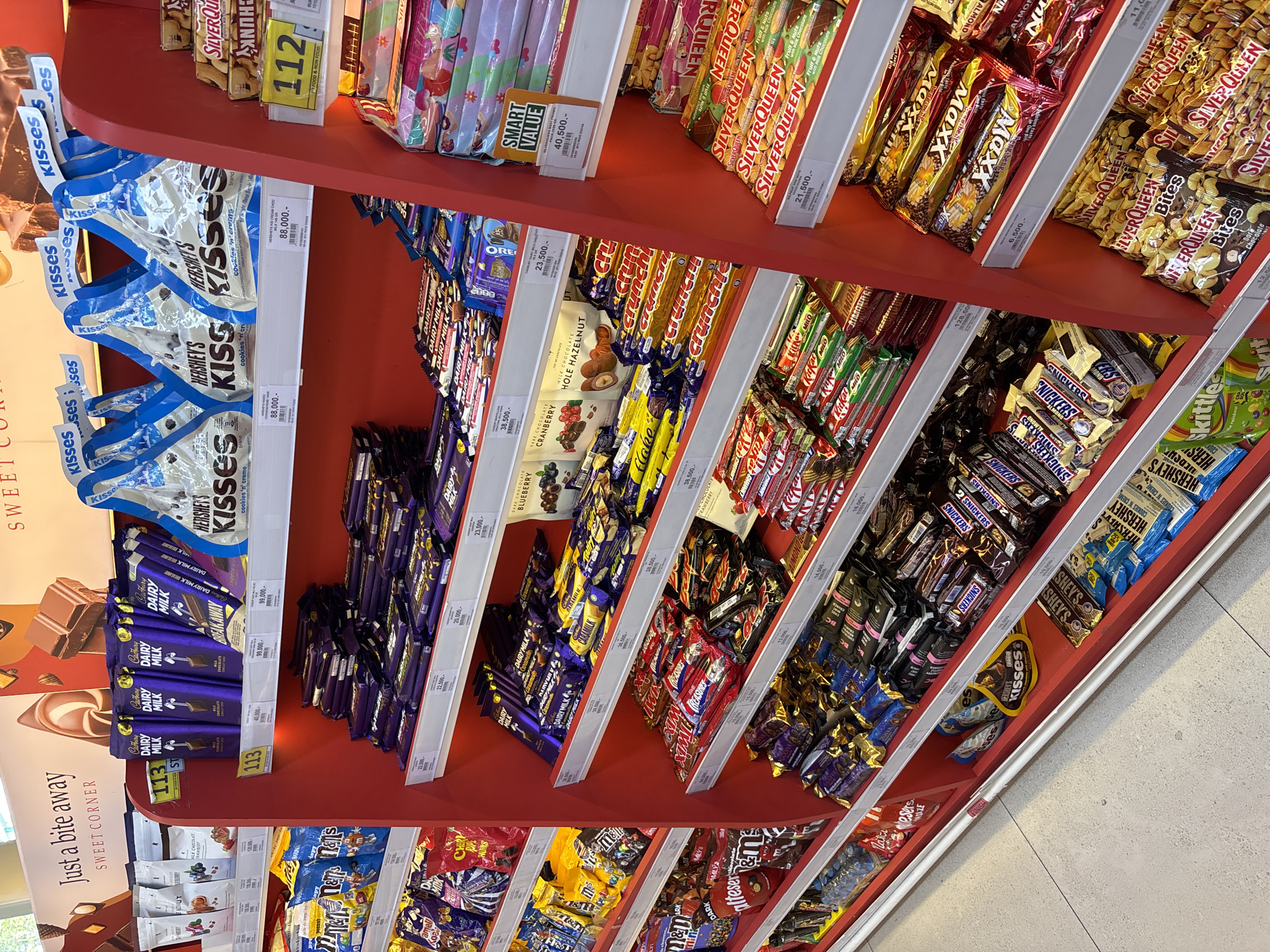 The image captures a retail display of chocolate products on shelves in Kuta Selatan, Indonesia. The shelves are predominantly red and are stocked with a variety of chocolate bars and candies. Brands visible include Hershey's Kisses, Cadbury Dairy Milk, SilverQueen, Snickers, and KitKat. Price tags accompany each product, indicating prices in Indonesian Rupiah. The bottom left corner of the image features signage with the text "Just a bite away SWEET CORNER".