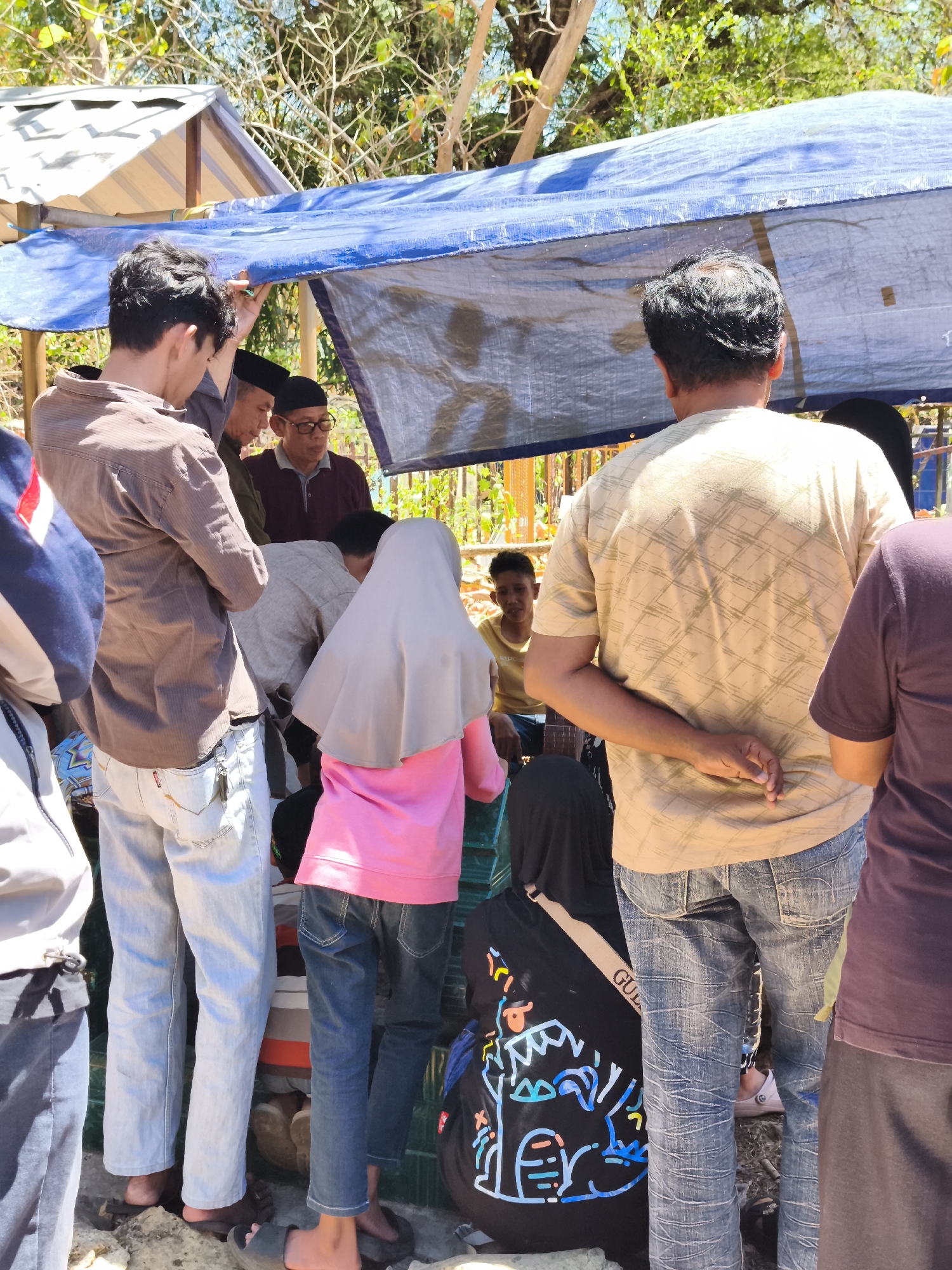 This image depicts a group of people gathered outdoors under a blue tarp. The scene appears to be in Tonasa, Indonesia, with lush green trees and vegetation visible in the background, suggesting a warm, tropical environment. Sunlight streams through the leaves, casting dappled shadows.
In the foreground, several individuals are visible from the back and side. A young man in a brown, striped shirt and light-wash jeans stands on the left, his hand resting on the tarp. Next to him, an older man in a maroon shirt and glasses, wearing a black kufi, is also present. Further into the scene, a person in a pink hijab and jeans is crouched or sitting. To the right, a man in a tan, patterned t-shirt and denim jeans stands with his hands clasped behind his back. Another person in a dark purple shirt is partially visible on the far right.
The ground is covered with rocks and debris. The blue tarp is supported by wooden poles or a frame, creating a makeshift shelter. The overall impression is of a casual gathering, possibly a community event or a market stall, taking place in a somewhat rustic outdoor setting in Tonasa, Indonesia.