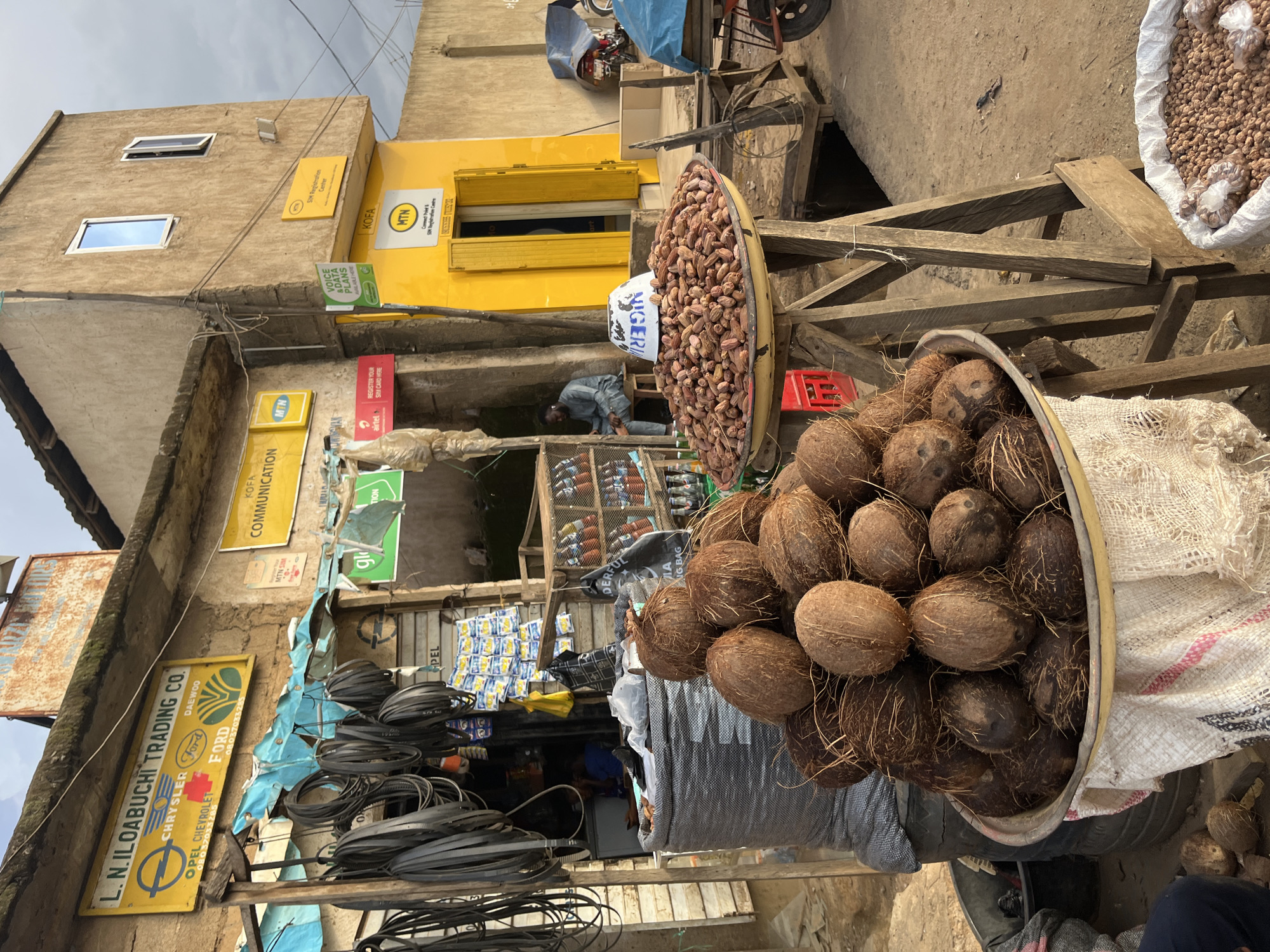 This image depicts a bustling street scene in Rafin Par, Nigeria. The foreground is dominated by a large basket overflowing with coconuts, some of which are spilling out onto a textured white sack and what appears to be a tire. To the left, a yellow sign with black lettering reads "L. N. ILOABUCHI TRADING CO." and lists various car brands like Chrysler, Opel, Chevrolet, Ford, and Daewoo, along with phone numbers. Above this, a sign for "MTN SIM REGISTRATION" is visible.
In the background, a bright yellow building with green shutters stands out. Several signs for telecommunications companies like MTN and Airtel are displayed on the building and surrounding walls, indicating a hub for mobile services. A person in a grey outfit is partially visible in the middle ground, seemingly engaged in some activity within a shop or stall. Various goods are displayed on shelves and in containers, including what looks like nuts in a large basket and packaged items. The overall atmosphere suggests a busy market or commercial area. The lighting and sky suggest it is daytime, likely overcast.