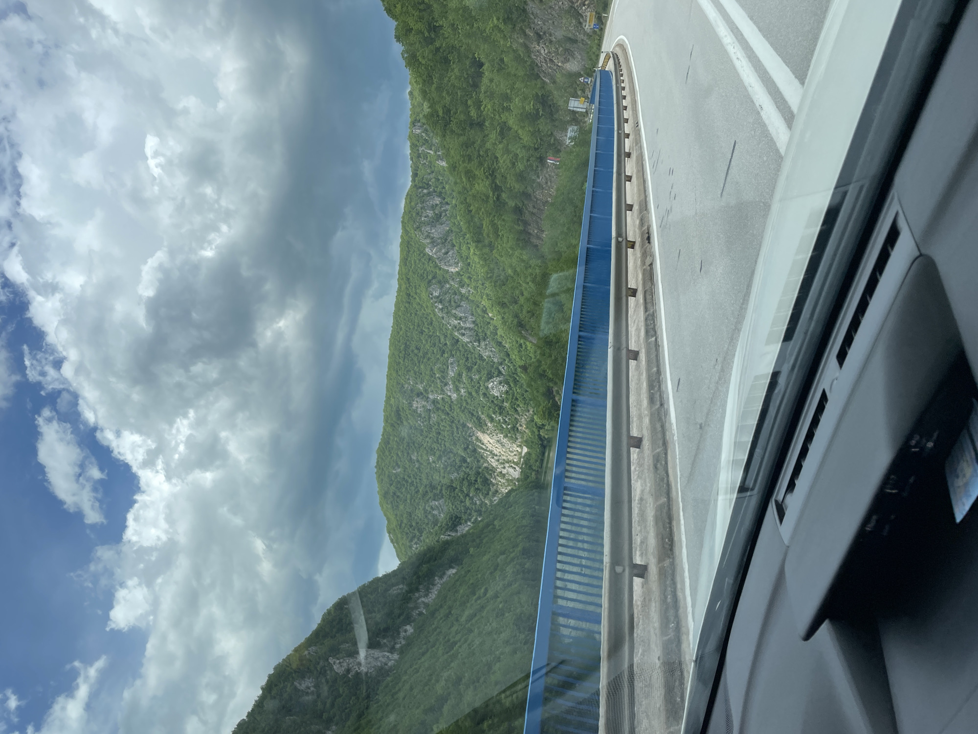 A view from inside a vehicle moving along a road in the Vlasinje area of Bosnia and Herzegovina. The vehicle is near a concrete barrier painted blue. A mountain covered in dense green foliage rises on the left, with a cloudy sky visible above. The road surface appears gray with white lane markings. The interior of the vehicle is partially visible, including the dashboard and some of the window frame.