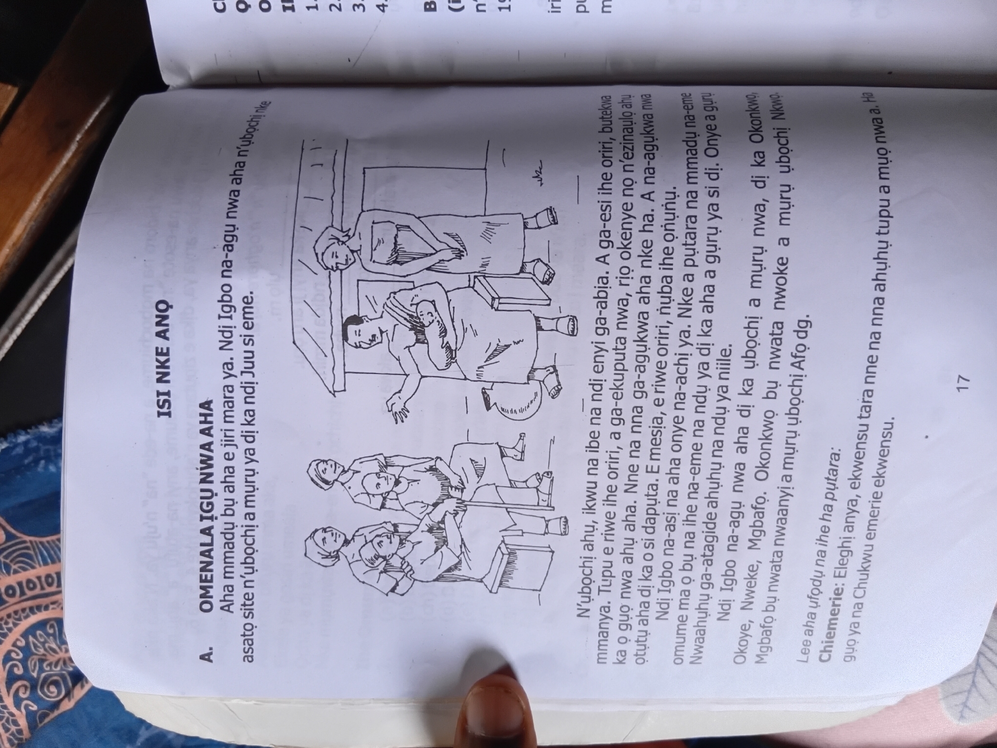 The image shows a page from a book with an illustration and text. The illustration depicts a group of people in what appears to be a village setting. On the left, three women are standing. The middle woman is holding a baby. In the center, two men are seated on a bench. On the right, a woman is standing next to a shelter. The text is in Igbo and discusses the "omenala igu nwa aha," which translates to "tradition of naming a child." The page is from a book with a numbered page at the bottom, "17." The upper right corner of the image shows a portion of another page from the same book with some text and numbers. Based on the visual context and the provided text, the scene is likely set in a traditional Igbo community. However, the specific location within Nkpor, Nigeria, cannot be determined from the image alone.