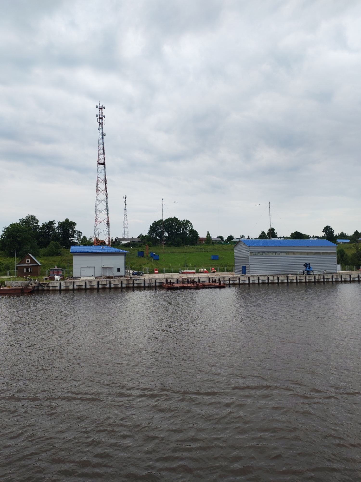 The image captures a scene at a waterfront in Ivanov Bor, Russia, on a cloudy day. In the foreground, a dark, rippling body of water dominates the lower half of the frame. Along the edge of the water is a concrete embankment or dock. On the embankment, several structures are visible.
To the left, there is a small, wooden, house-like building with a pitched roof and a single window. Next to it are some red and white buoys and industrial equipment. Further to the right, a larger, modern-looking industrial building with a blue, corrugated metal roof and gray walls stands prominently. This building has a large garage-style door and a smaller entrance door. Adjacent to this building is some blue machinery.
In the background, a tall, lattice-like radio tower is the most striking feature, with several antennas mounted at different levels. Another, shorter radio tower is visible behind the larger industrial building, and a third, even smaller tower is seen in the distance. The land behind the structures is a green, grassy slope dotted with trees. A few other smaller buildings and what appear to be shipping containers are scattered across the landscape. The sky is overcast with gray clouds. Several barges or flat-bottomed boats are moored along the dock. There are no people visible in the image.