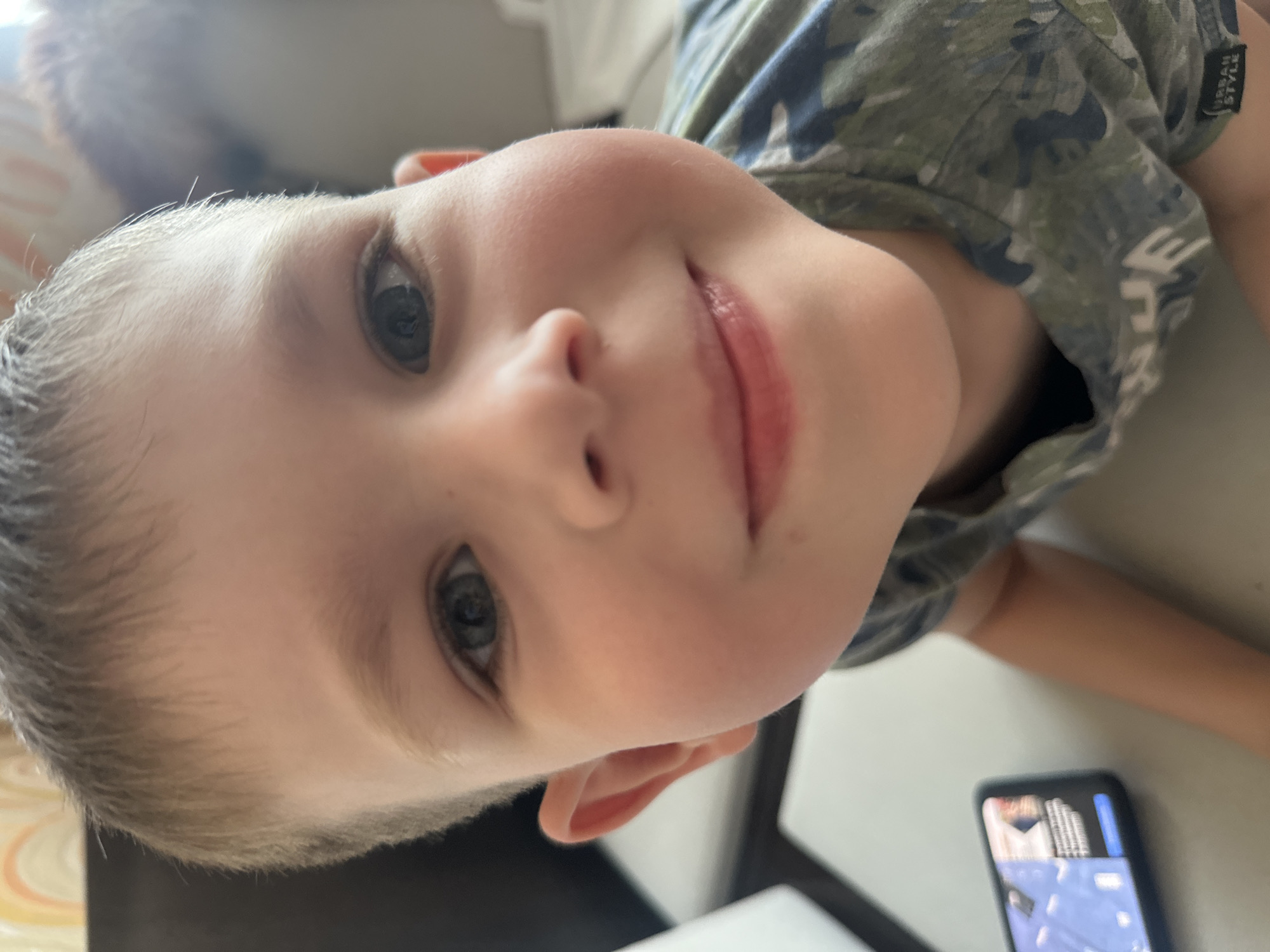 A close-up, eye-level, medium shot captures the face of a young boy, likely Caucasian, with short brown hair and blue eyes. He is looking directly at the camera with a slight smile. The boy is wearing a camouflage t-shirt. To the left of the boy, a smartphone is visible, displaying a screen with what appears to be a game. The background is blurred, showing hints of furniture and a patterned wall, suggesting an indoor setting. The overall lighting is bright, indicating daylight. The scene is consistent with a casual home environment in Novoaltaysk, Russia.
