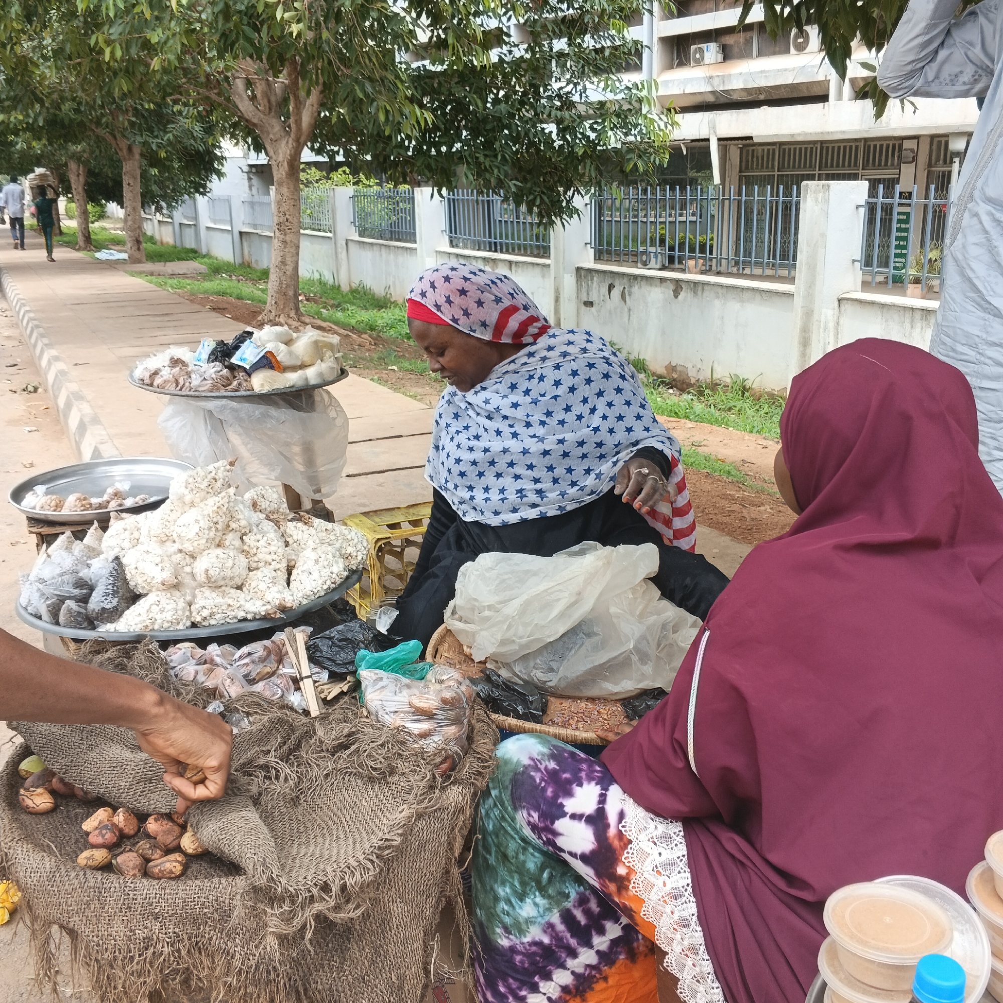 The image depicts an outdoor market scene in Shaba, Nigeria. Two women are present, one wearing a headscarf patterned with stars and stripes, and the other, a woman in a maroon headscarf, faces away from the camera. They are surrounded by various food items displayed on metal trays and burlap sacks. The foreground shows a hand reaching into a burlap sack filled with nuts. In the background, a row of trees lines a sidewalk, with a building and a fence visible behind them. The overall setting appears to be a street-side vendor stall.