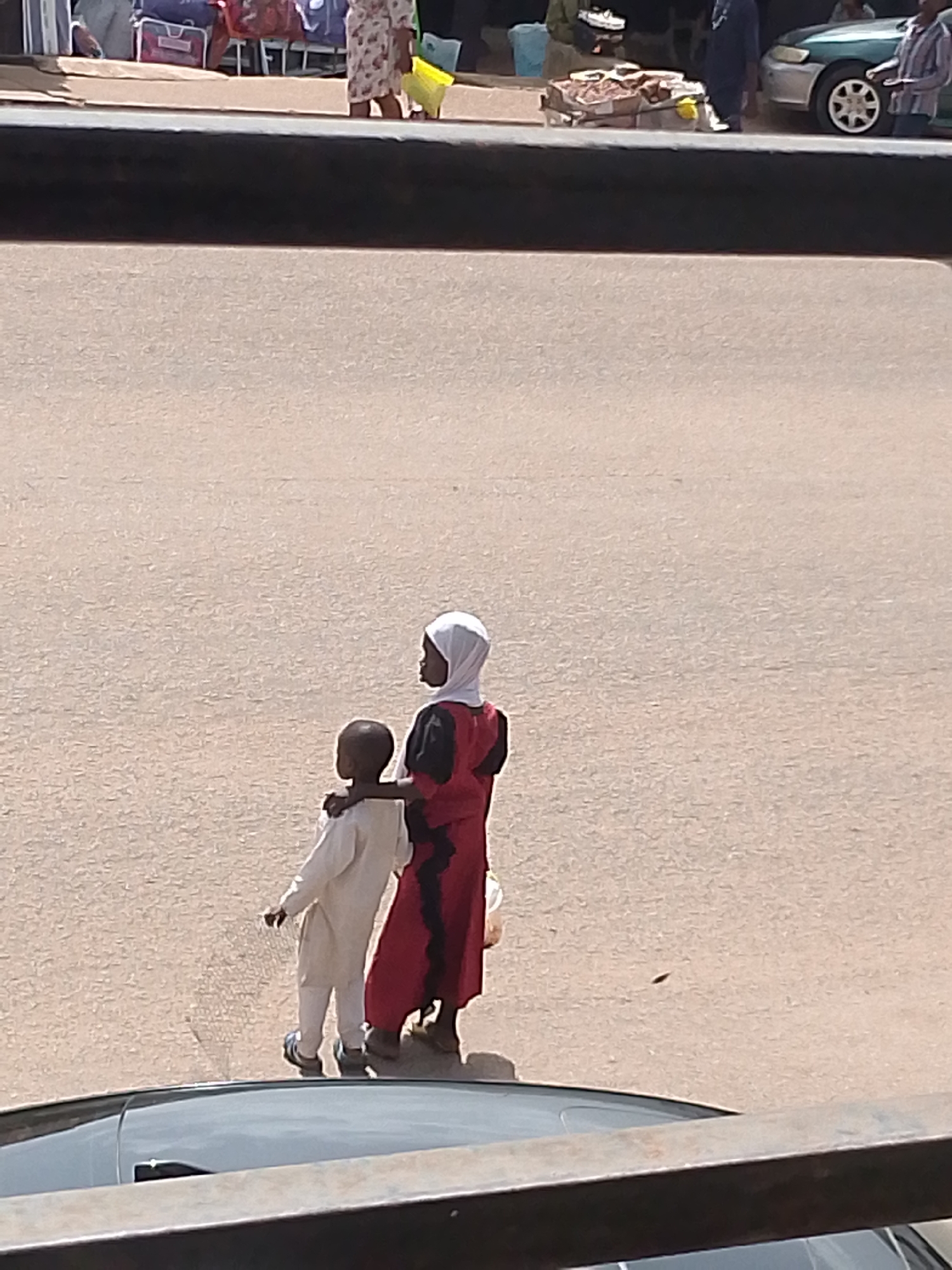 In Bukuru, Nigeria, two young individuals, one appearing to be a child and the other slightly older, stand on a dusty, paved surface, possibly a street or marketplace. The elder, wearing a red garment and a white headscarf, has their arm around the shoulders of the younger individual, who is dressed in a light-colored outfit. In the background, blurred figures and merchandise suggest a bustling area, with glimpses of a car and other vehicles hinting at urban activity. The overall impression is of a moment captured in a vibrant, lively setting, with the two individuals sharing a quiet connection amidst the surrounding scene.