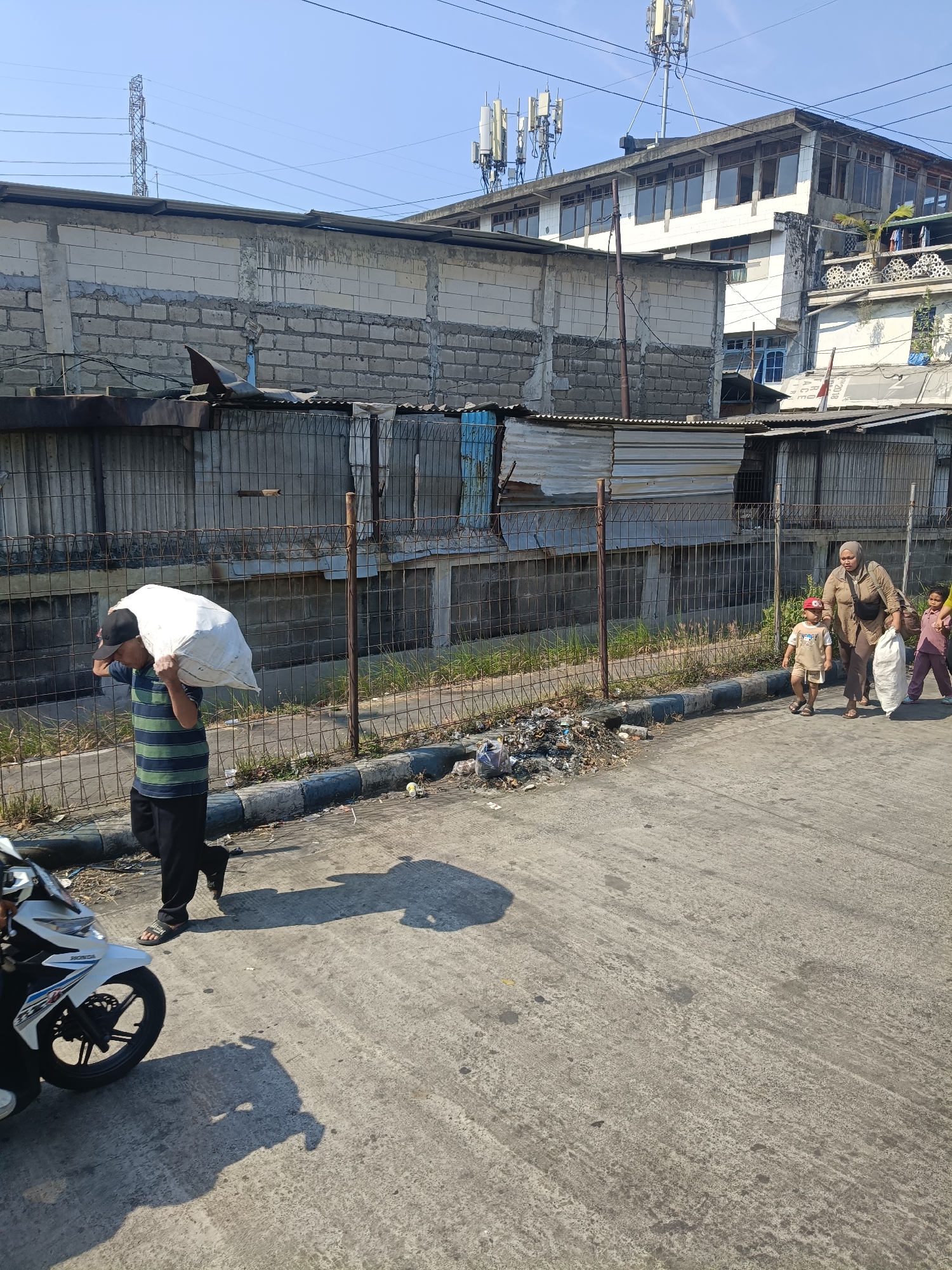In Kedungrejo, Indonesia, a man is walking on a paved street carrying a large white sack over his right shoulder. He wears a black baseball cap, a horizontally striped green and blue shirt, and black pants. He is walking on the left side of the street, and a white motorcycle is partially visible in the lower left corner. Further down the street, a woman and two children are walking from right to left. The woman wears a brown top and dark pants and carries a white sack. The child in front wears a red helmet, a light-colored shirt, and shorts. The child behind wears a pink shirt and light-colored pants. A fence runs along the right side of the street, separating it from a grassy area and structures that appear to be made of cinder blocks and corrugated metal. Piles of trash are visible on the side of the road near the fence. In the background, buildings with multiple windows and cellular towers are visible against a clear blue sky.