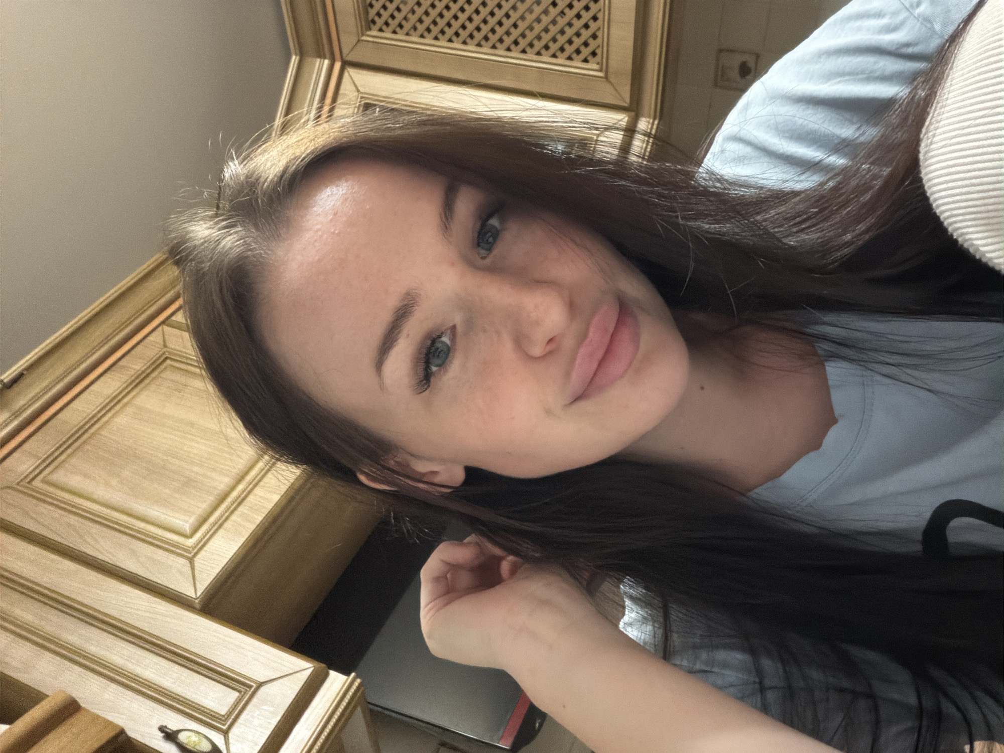 A young woman with long, dark brown hair and light-colored eyes (likely blue or green) is captured in a close-up, angled selfie. She has a fair complexion with visible freckles across her nose and cheeks and displays a subtle, closed-lip smile while looking at the camera. She wears a light blue top, partially visible, and her right hand is seen in the lower part of the frame, seemingly resting near her face or hair.
The background features ornate wooden cabinetry with a light stain and decorative paneling, characteristic of a well-furnished indoor space, possibly a kitchen or home setting. The image is taken from a low, upward-looking perspective, resulting in a tilted composition. On the right, a light-colored, possibly ribbed, fabric is visible. A small golden object with green detailing rests on a lower wooden surface, and a dark rectangular object is also present. The lighting is soft and ambient, indicative of an indoor environment. No text is visible within the image. The setting is consistent with a home interior, and the provided context indicates the location is Russia. The woman appears relaxed and content.