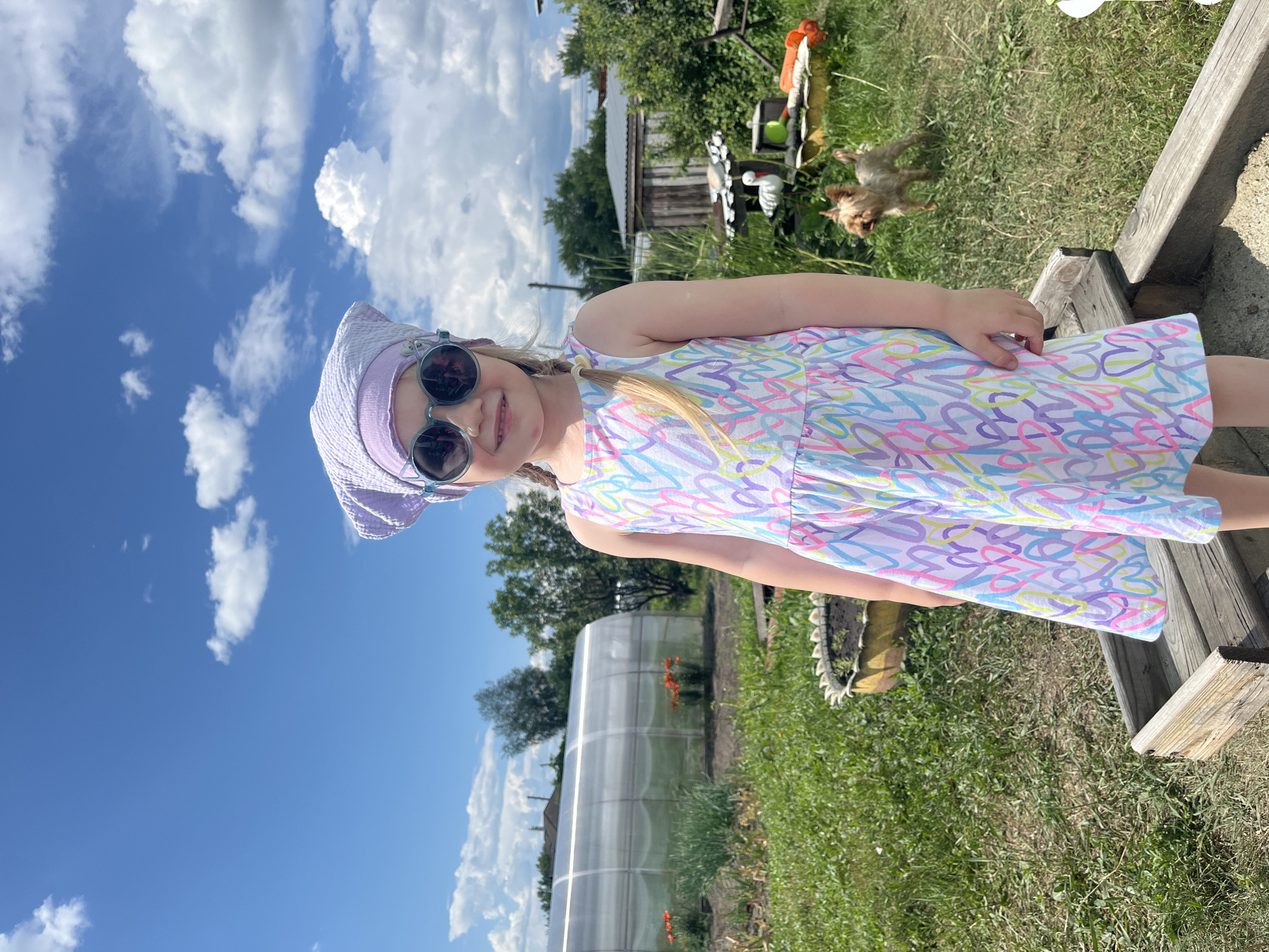 A young girl with blonde pigtails and a light purple headscarf is standing in a sandbox outdoors. She is wearing sunglasses and a sleeveless dress with a pastel heart pattern. In the background, a greenhouse, a small wooden building, trees, and a small dog are visible. The sky is blue with white clouds. The location appears to be a rural setting, possibly a backyard or garden in Сухринское, Russia.
