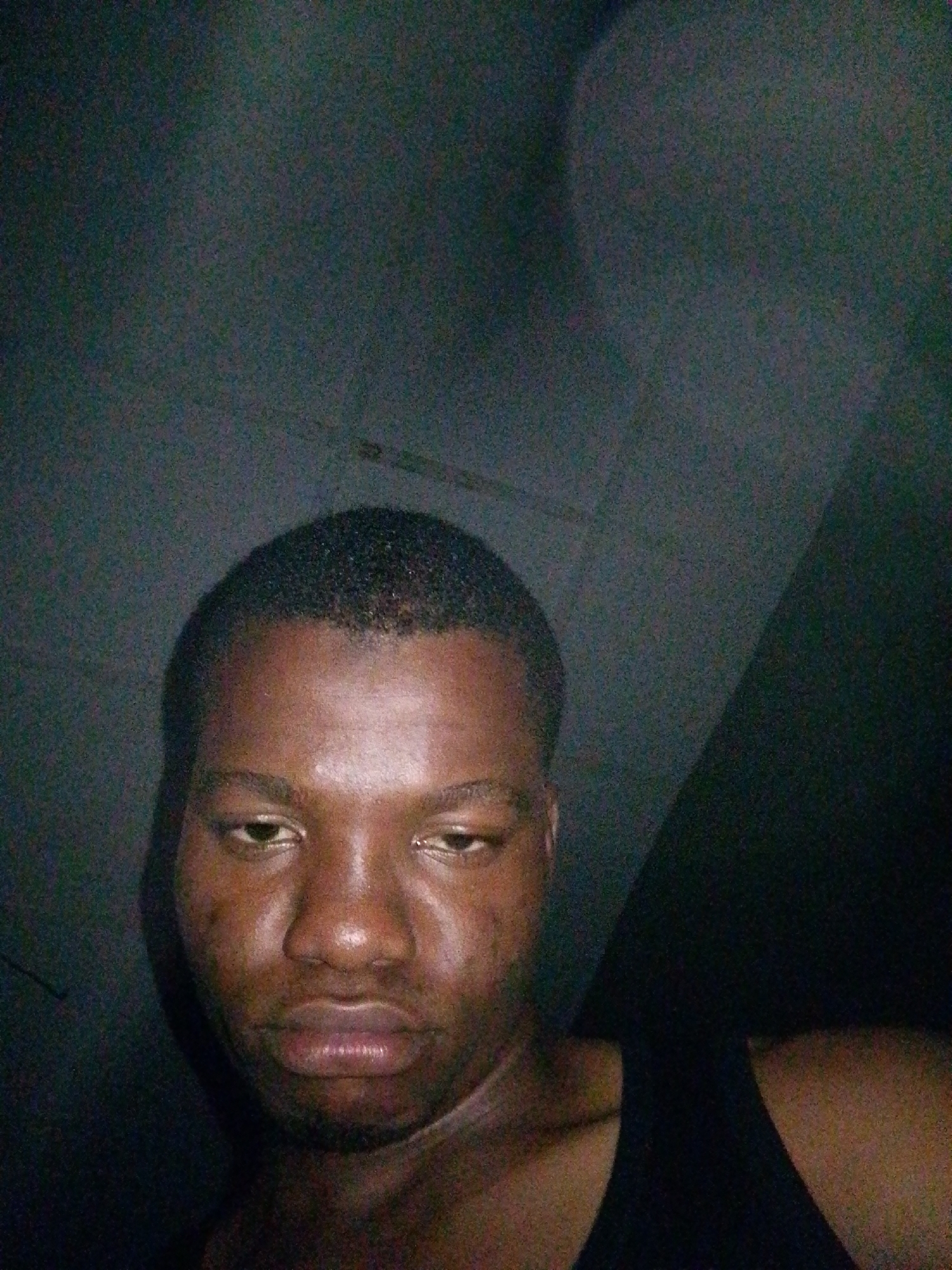 The image is a close-up, low-light selfie of a Black man. He is wearing a dark tank top. The background appears to be a dark, textured wall or ceiling, possibly in a domestic setting in Ipetumodu, Nigeria. The lighting is dim, creating shadows and highlighting the subject's face, particularly his forehead, nose, and lips. His expression is somewhat somber. The image quality is grainy due to the low light conditions.