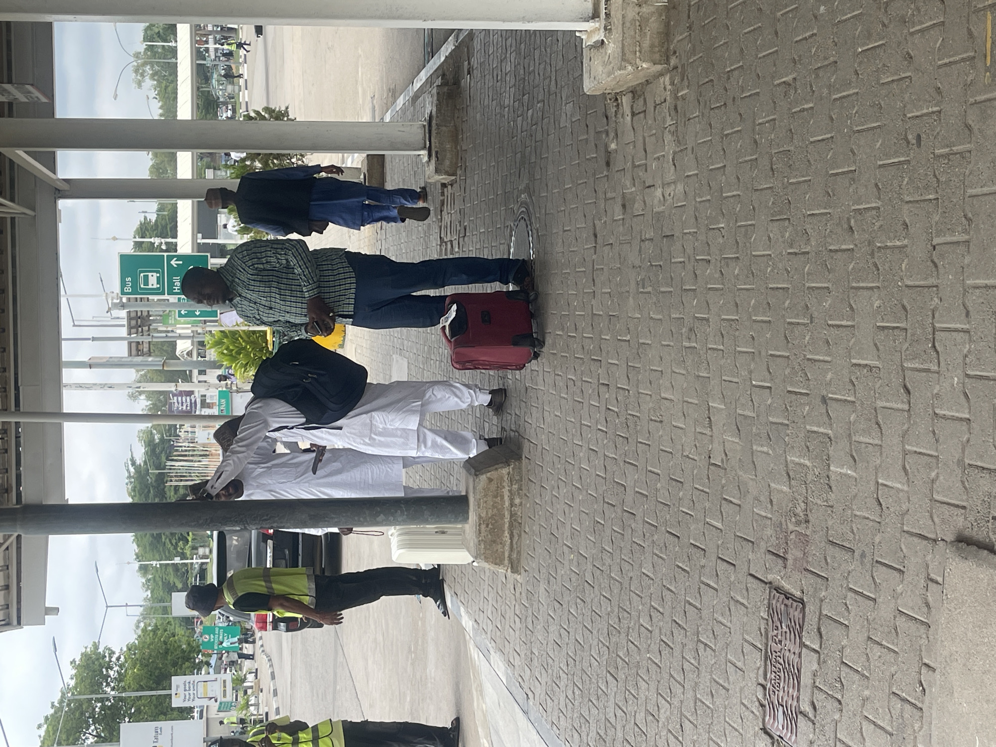 The image depicts an outdoor covered waiting area, likely at a transportation hub such as an airport or bus terminal in Gwaska, Nigeria, during daylight hours. The sky appears bright and overcast.
In the foreground, the ground is paved with grey interlocking concrete blocks. Several individuals are standing or waiting. Towards the center-left, a man wearing a green and white plaid shirt and dark blue jeans stands beside a red rolling suitcase, holding a mobile phone. Behind him, another man in a blue tunic and cap is visible. Further to the left, a man dressed in white traditional attire (possibly a thobe or kaftan) with a black backpack stands near a white rolling suitcase. He appears to be looking down or speaking to someone. Two individuals wearing yellow high-visibility vests and dark trousers, possibly security or staff, are positioned on the far left and right side of the foreground and midground.
The area is covered by an overhead structure composed of concrete and metal beams, providing shade. In the background, a roadway is visible with several parked and moving vehicles, including a white van. Greenery, consisting of trees and shrubs, dots the landscape beyond the road.
Notable text includes a green road sign displaying a bus icon next to "Bus" with a left-pointing arrow, and "Hall" with an upward-pointing arrow. Another yellow sign in the distance is partially visible, showing "Your goods. Your smile." and "Tatum Bank". A manhole cover embedded in the pavement in the midground has the text "FEDERAL ROADS" partially visible. The overall scene suggests a routine day at a busy public transport facility.