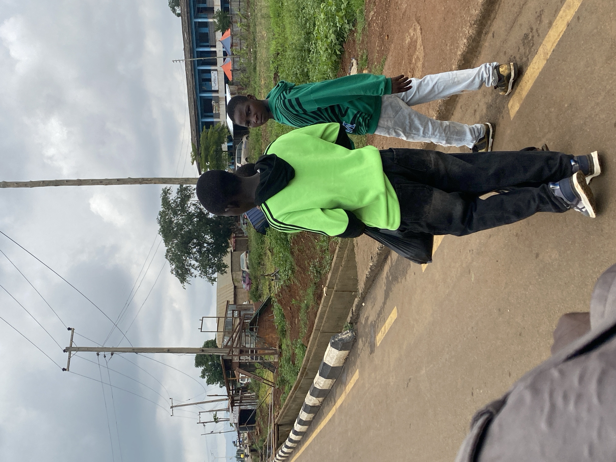 Two youths are standing on a paved road with a yellow line marking. One wears a green hoodie with black stripes and black pants. The other wears a green sweatshirt with black patterns and light-colored pants. A utility pole and wires are visible in the background, along with buildings and vegetation. The location is likely Kafanchan B, Nigeria, as per the prompt. A white and black striped barrier runs along the edge of the road.