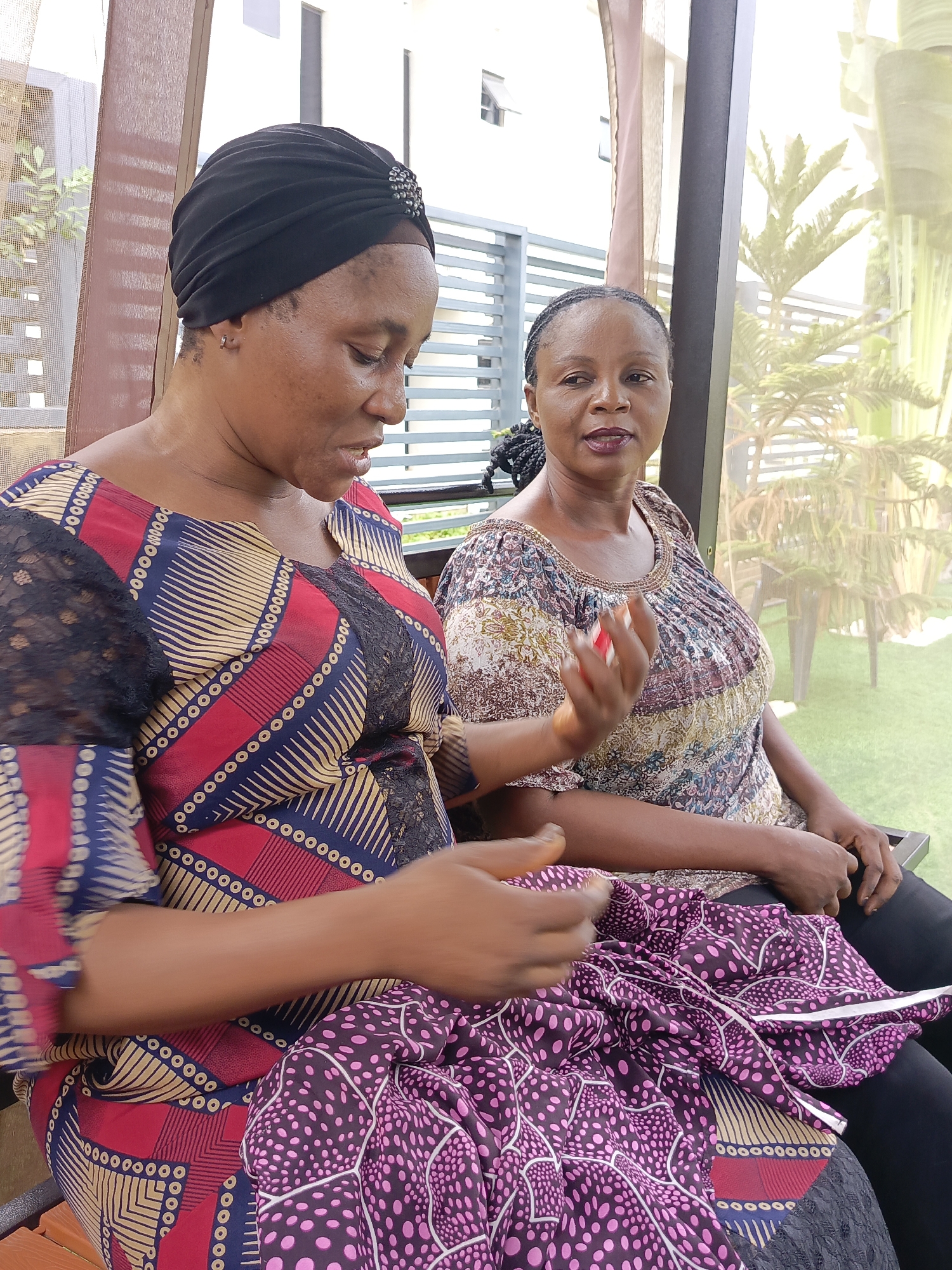 The image captures two women seated outdoors in a bright, daytime setting, likely in Akwanga, Nigeria. In the foreground, a woman with a dark complexion wears a black turban adorned with a beaded embellishment and a vibrant dress featuring red, blue, and yellow geometric patterns with black lace sleeves. She is looking downwards, intently focused on a piece of purple fabric with pink polka dots in her lap, suggesting she is engaged in sewing or a similar handiwork.
Behind her, a second woman, also with a dark complexion, is seated. She wears a colorful, intricately patterned top with a round neckline and has her hair styled in braids. She looks towards the camera with a calm, neutral expression, holding a small red object in her right hand.
The setting appears to be an outdoor patio or veranda, with a dark wooden support beam visible. The background reveals lush tropical greenery, including tall, palm-like plants and other foliage, indicative of a garden environment. A modern white building with visible windows can be seen further back, and the ground is covered with what appears to be artificial green grass. The scene portrays a casual, relaxed moment of quiet activity and companionship under clear daylight conditions. No visible text is present.