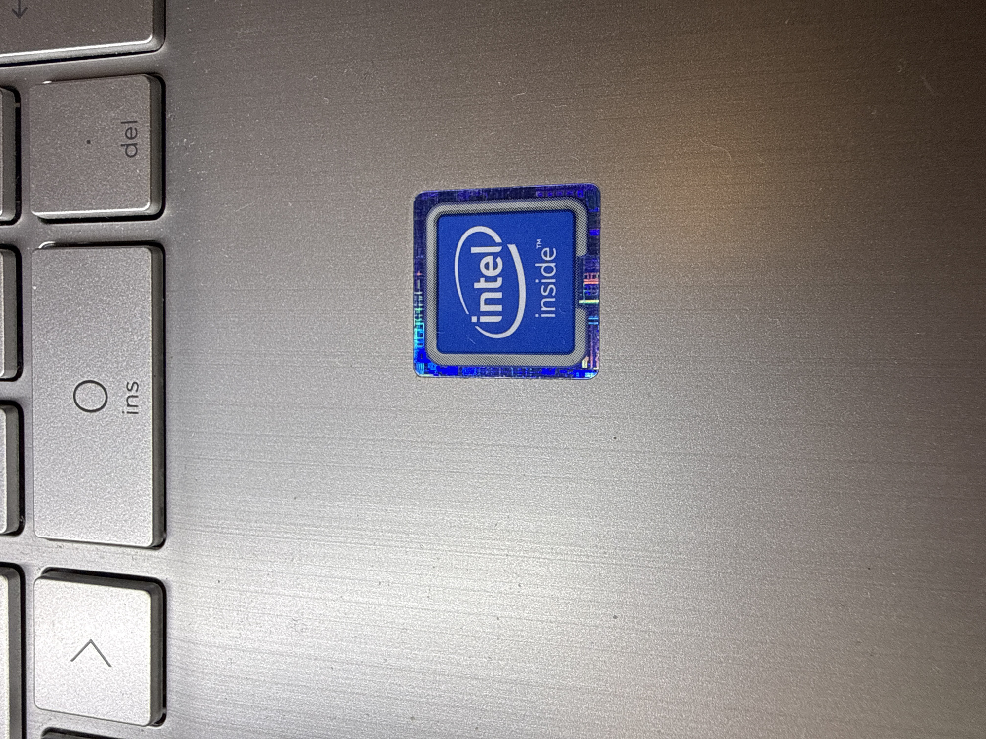 A close-up, overhead shot shows the keyboard and an Intel sticker on a silver laptop. The Intel sticker, located to the right of the center of the frame, has a blue background with the "Intel Inside" logo. Several keys are visible, including the right arrow, "ins," and "del" keys. The laptop's surface has a brushed metal texture.