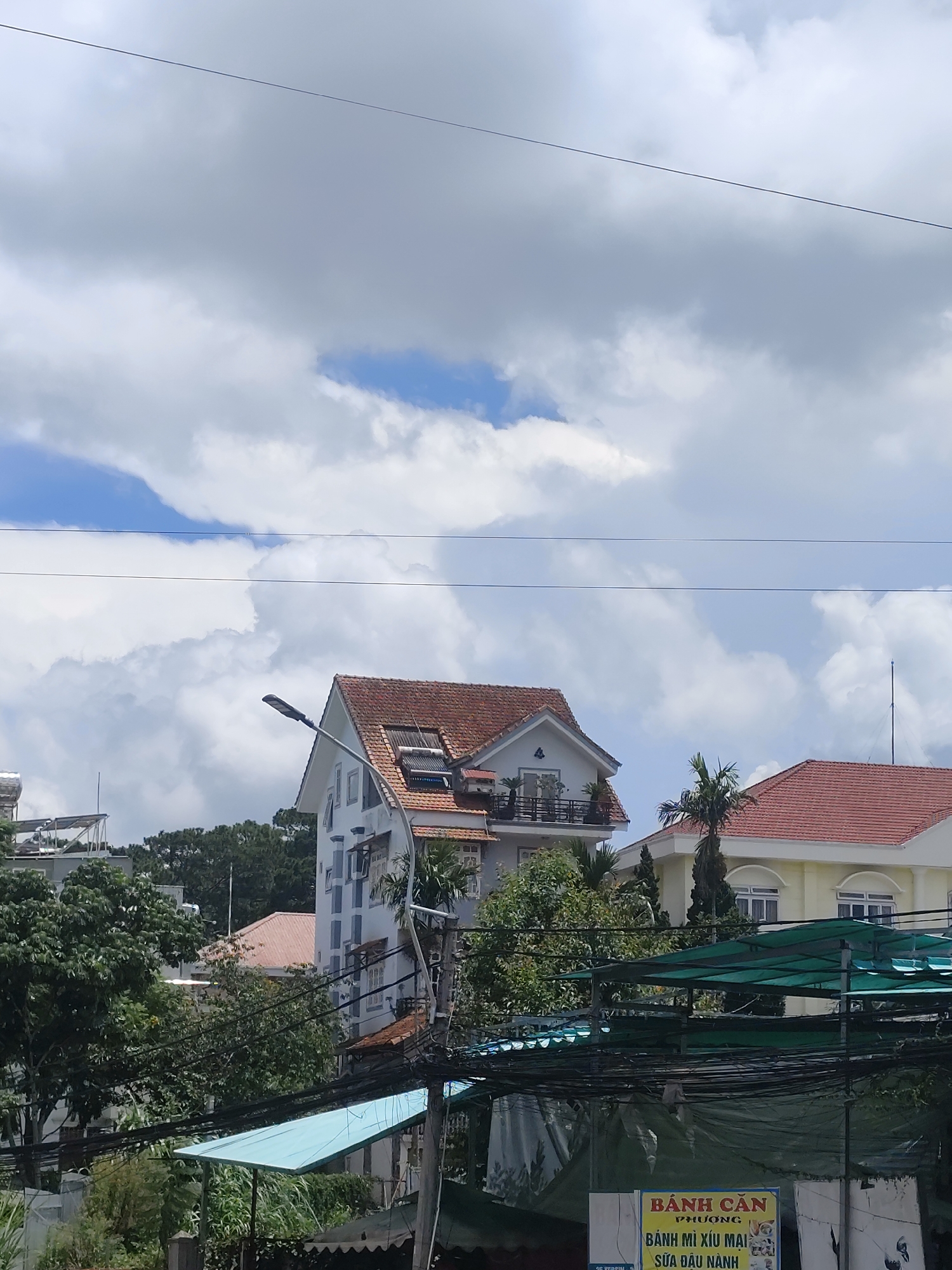 The image depicts a view of residential buildings and trees against a cloudy sky in Da Lat, Vietnam. The central focus is a multi-story white house with a reddish-brown tiled roof, featuring multiple windows and balconies. To the right of this house is another building with a similar tiled roof and yellow exterior. In the foreground, there are power lines, a blue awning, and lush green trees. Further in the background, more trees and buildings are visible, suggesting a dense urban or semi-urban environment. The sky is filled with large, fluffy white clouds interspersed with patches of blue sky. A sign with Vietnamese text, possibly advertising food, is partially visible at the bottom right of the frame. The overall scene conveys a typical day in a Vietnamese city with its characteristic mix of architecture, vegetation, and infrastructure.