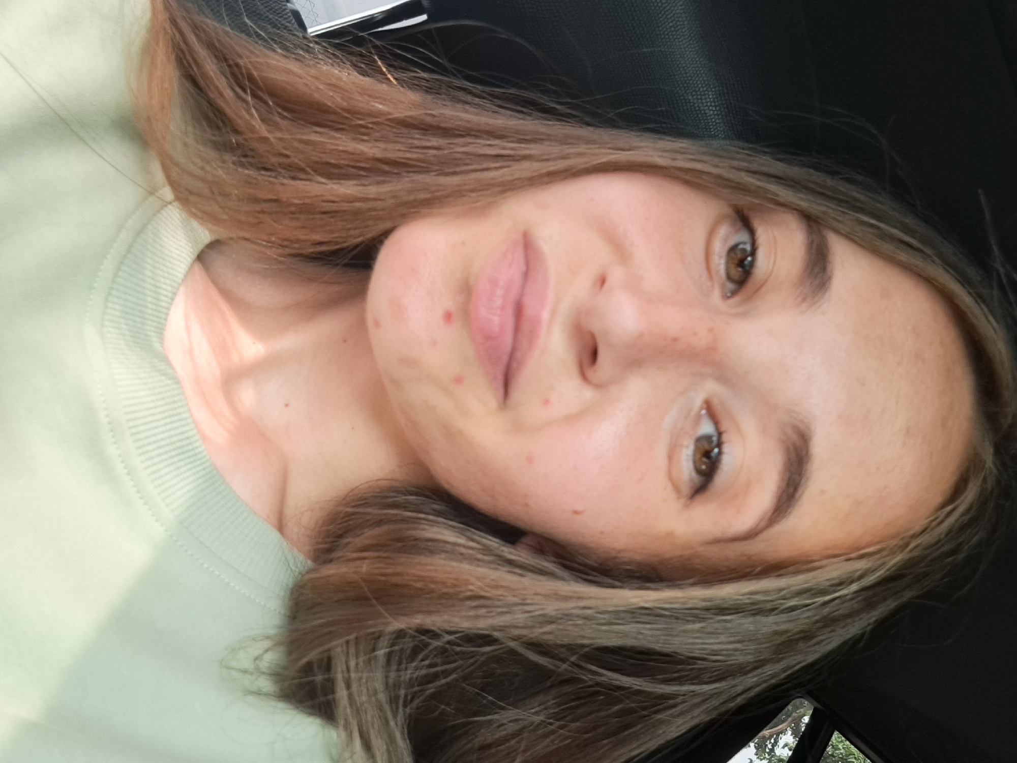 The image shows a young woman with light brown hair and brown eyes, looking at the camera. She is wearing a light green sweatshirt. Her face has several freckles and a few blemishes. The background is dark, suggesting she may be in a car. There are no discernible landmarks or architectural features visible that would indicate a specific location within Vypasne, Ukraine. The lighting suggests it is daytime.