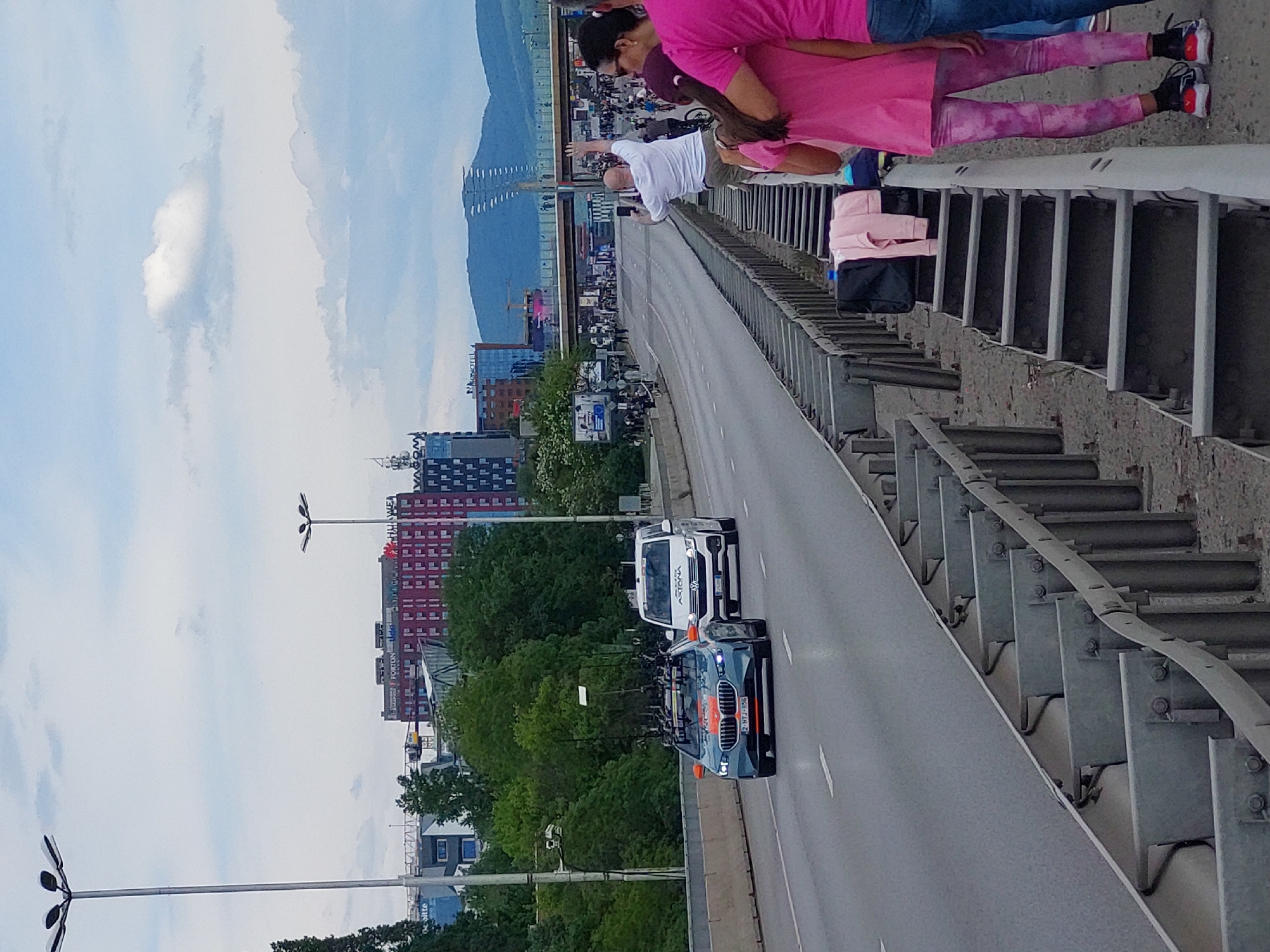 The image shows a view of a bridge in Sofia, Bulgaria, likely the Stamboliiski Bridge. The roadway is visible, with a dark car and a white van driving on it. A crowd of people stands on the sidewalk, looking towards the road. Buildings can be seen in the background, including one with red and white markings. Mountains rise in the distance under a cloudy sky. The bridge's metal railings are prominent in the foreground.