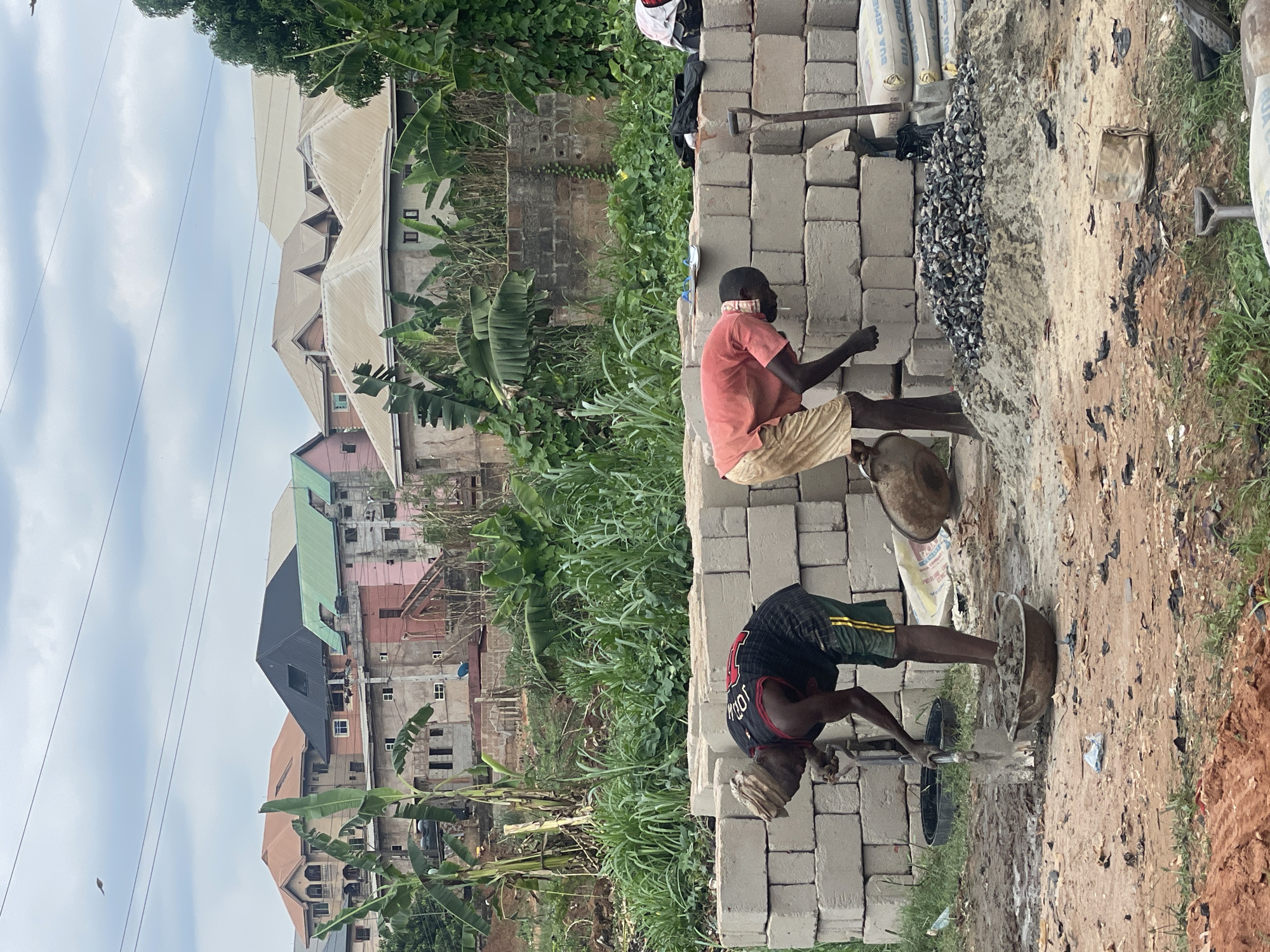 The image captures an outdoor construction scene in Obosi, Nigeria, during daytime under a partly cloudy sky. Two men are actively engaged in manual labor, likely preparing building materials.
In the foreground, large stacks of gray concrete blocks are prominent, alongside piles of sand and dark gravel on the muddy ground. Several sacks of cement are visible, with one clearly displaying the text "BUA CEMENT." Various tools, including shovels and a pickaxe-like implement, are scattered nearby.
One man, dressed in a red/orange t-shirt and light-colored shorts, is seated on a stack of concrete blocks. He appears focused on handling materials in a metal pot on the ground, with a small, light-colored object in his mouth. The second man, wearing a dark sleeveless top (possibly a jersey with "JOHN" visible on the back) and dark shorts with yellow stripes, is bent over, actively mixing materials in a large metal bowl on the ground using a tool.
The background reveals a densely packed residential area with numerous buildings of varying sizes and colors, many featuring corrugated metal roofs. Some structures appear to be unfinished or under construction. Lush green vegetation, including banana plants and tall grass, grows abundantly throughout the scene. Power lines traverse the sky, and a small bird is visible flying in the upper left. The overall scene depicts ongoing manual construction work in a vibrant, developing community.