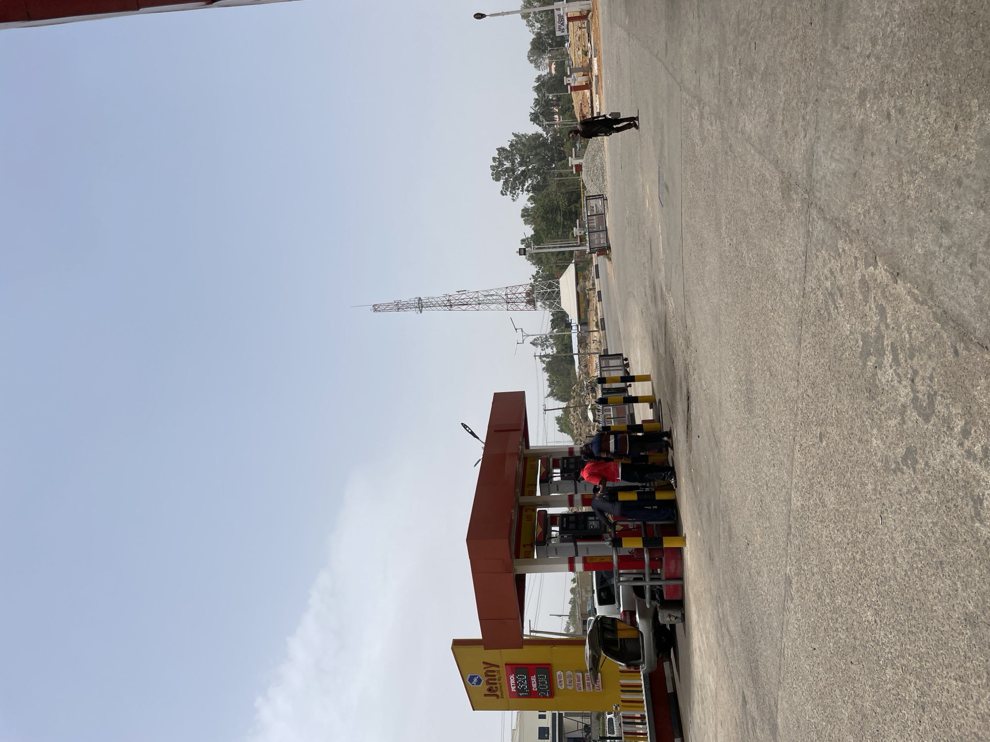 The image depicts a gas station under a clear sky. The station, branded "Jenny Investment Nig Ltd", displays fuel prices: Petrol at 1.320 and Diesel at 2.000. At the pumps, several individuals in dark clothing are present, and one is wearing a red shirt. Behind the station, a lattice-style tower rises. The scene is situated on a wide paved area with a person walking in the distance. Based on the brand name and visual clues, the location is possibly in Jos, Nigeria.