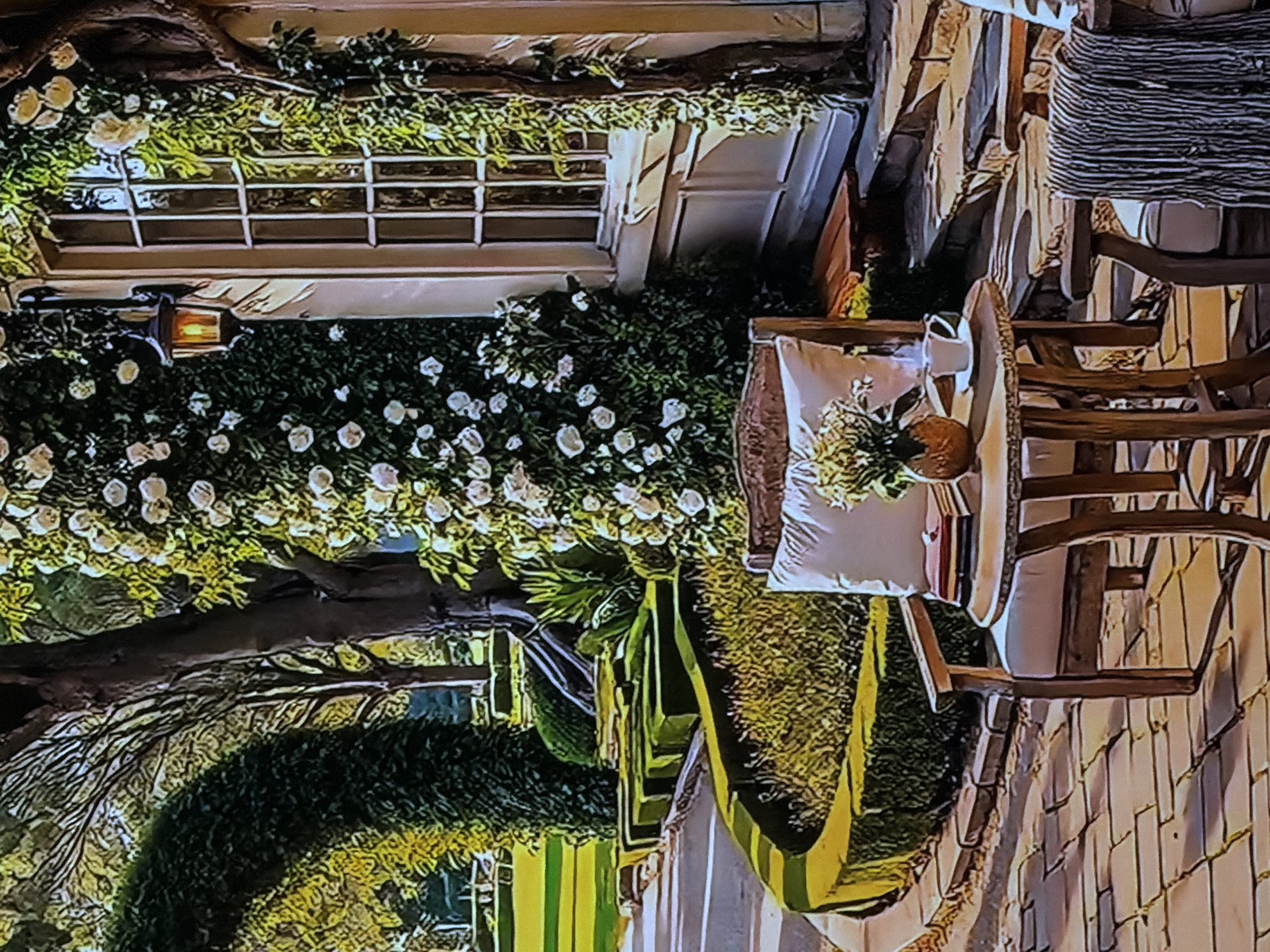 This media file captures an elaborately decorated outdoor setting, likely prepared for a special event such as a wedding or garden party, in Sofiivska Borshchahivka, Ukraine. The scene is illuminated with a warm glow, suggesting it is either evening or night, or an indoor setting with atmospheric lighting.
The central focus is a white wooden window frame, richly adorned with lush green foliage and numerous white flowers, possibly roses, creating a romantic and ethereal ambiance. A vintage-style lantern with a bright, warm light hangs below the window, casting a soft glow.
To the right of the decorated window, a cozy seating area is arranged with two ornate wooden chairs. One chair features a white cushion adorned with a small floral arrangement and a decorative brown gourd or pumpkin. The other chair has a textured, possibly woven, blanket or throw draped over its back. The ground beneath the chairs appears to be a light-colored paved or planked surface.
Further to the left, a large tree trunk with intertwining bare branches and green vines is visible, adding to the natural, garden-like atmosphere. In the background, there's more dense green foliage, possibly a manicured hedge or topiary, and a distinctive green and yellow striped object lies on the ground, perhaps a rug or mat. No people or visible text are present in the image.
