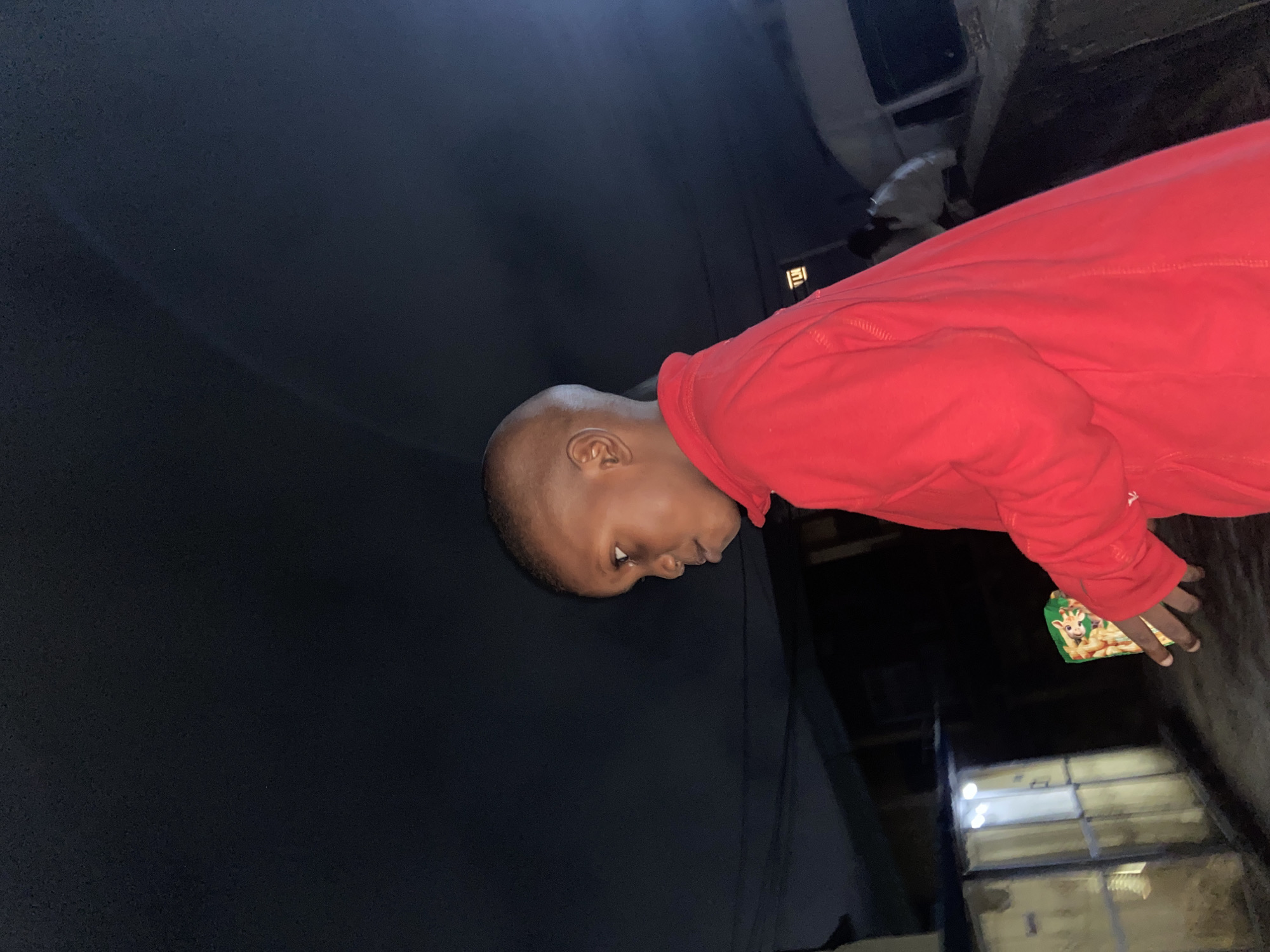 A young boy with a buzz cut, wearing a bright red long-sleeved shirt, is captured in a medium shot. He is positioned in the foreground, facing to the left of the frame, with his profile visible. His gaze is directed downwards and slightly to the left. In his right hand, he holds a small, colorful box, featuring an illustration of a giraffe.
The background suggests an urban environment at night. To the left, a building with illuminated windows and a metallic-looking roof or awning is visible. Further back, a white vehicle, possibly a van or bus, is present, with a person in a light-colored garment partially visible behind it. The overall lighting is dim, with the primary light source appearing to be from the building's windows. The sky is dark and appears to be overcast or obscured. The ground surface is rough and uneven, suggesting an unpaved or distressed road or pathway. The scene gives the impression of being in a less developed part of a city, possibly on the outskirts. The image was taken at night in the city of Lagos, Nigeria.