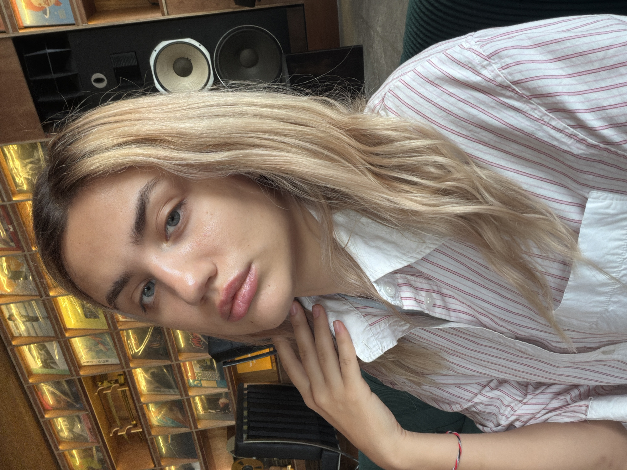 A young Caucasian woman with blonde hair and blue eyes is photographed from the chest up, looking towards the camera. She is wearing a white and red striped button-up shirt. Her left arm is raised, with her hand resting on her neck. In the background, shelves are filled with vinyl records. To the right of the woman, a large black speaker is visible. The scene appears to be indoors, possibly in a cafe or shop. There is no discernible connection to North Kuta, Indonesia.