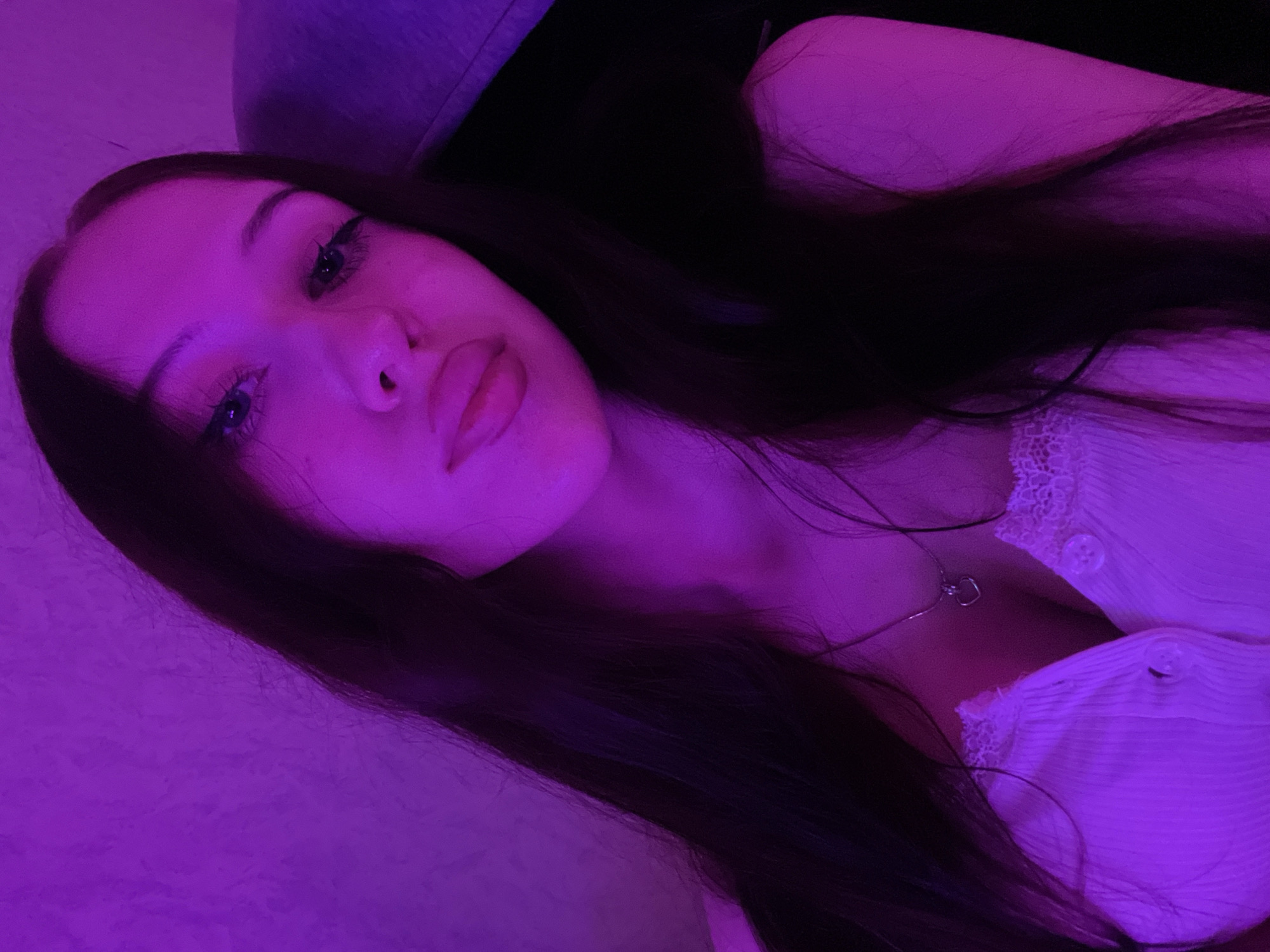 The image is a close-up, indoor selfie of a young woman with long, dark hair. She is illuminated by purple lighting, which casts a pinkish-purple hue across her skin and clothing. Her eyes are dark and her lips are full. She is wearing a white top with lace trim and a delicate necklace with a heart pendant. The background appears to be a textured wall, also lit with purple light. The overall mood is intimate and atmospheric. There is no information in the image to suggest a location in Votkinsk, Russia.