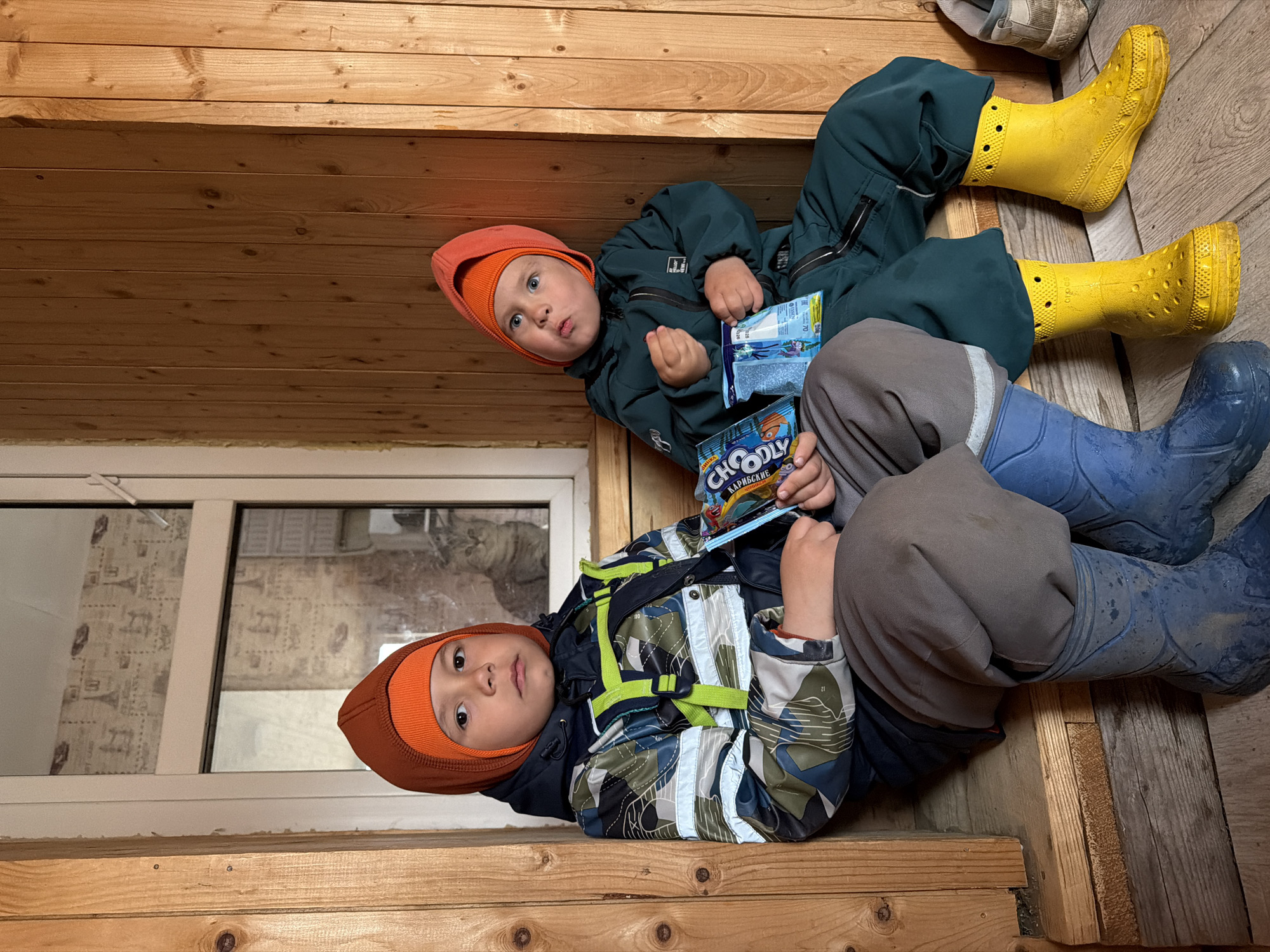 Two young children are lying on a wooden surface. The child on the left is wearing a camouflage jacket with neon green straps, grey trousers, and blue rubber boots. He holds a "Choodly" candy bar. The child on the right wears a green jumpsuit and yellow rubber boots, holding another candy bar. Both children wear orange balaclavas. A window is visible in the background with patterned curtains. The setting appears to be indoors, with wooden walls and flooring. The candy bar wrapper indicates the location is likely Chaikovsky, Russia.