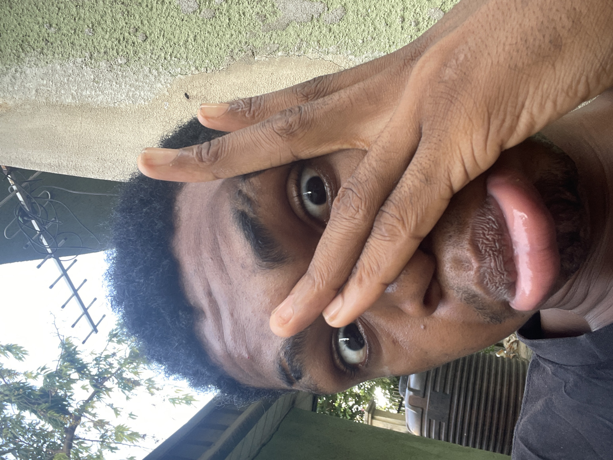 A close-up, slightly rotated image depicts a man with dark skin and curly black hair sticking his tongue out. His hand partially covers his face, with his fingers spread across his forehead and eye. The man’s eyes are wide and expressive. He is wearing a dark shirt. The background shows a textured wall, possibly concrete, and some greenery. There is an antenna and a tree in the distance, potentially indicating an outdoor setting. While the image shows visual elements, there is not enough information to pinpoint the location in the city of Ado, Nigeria.