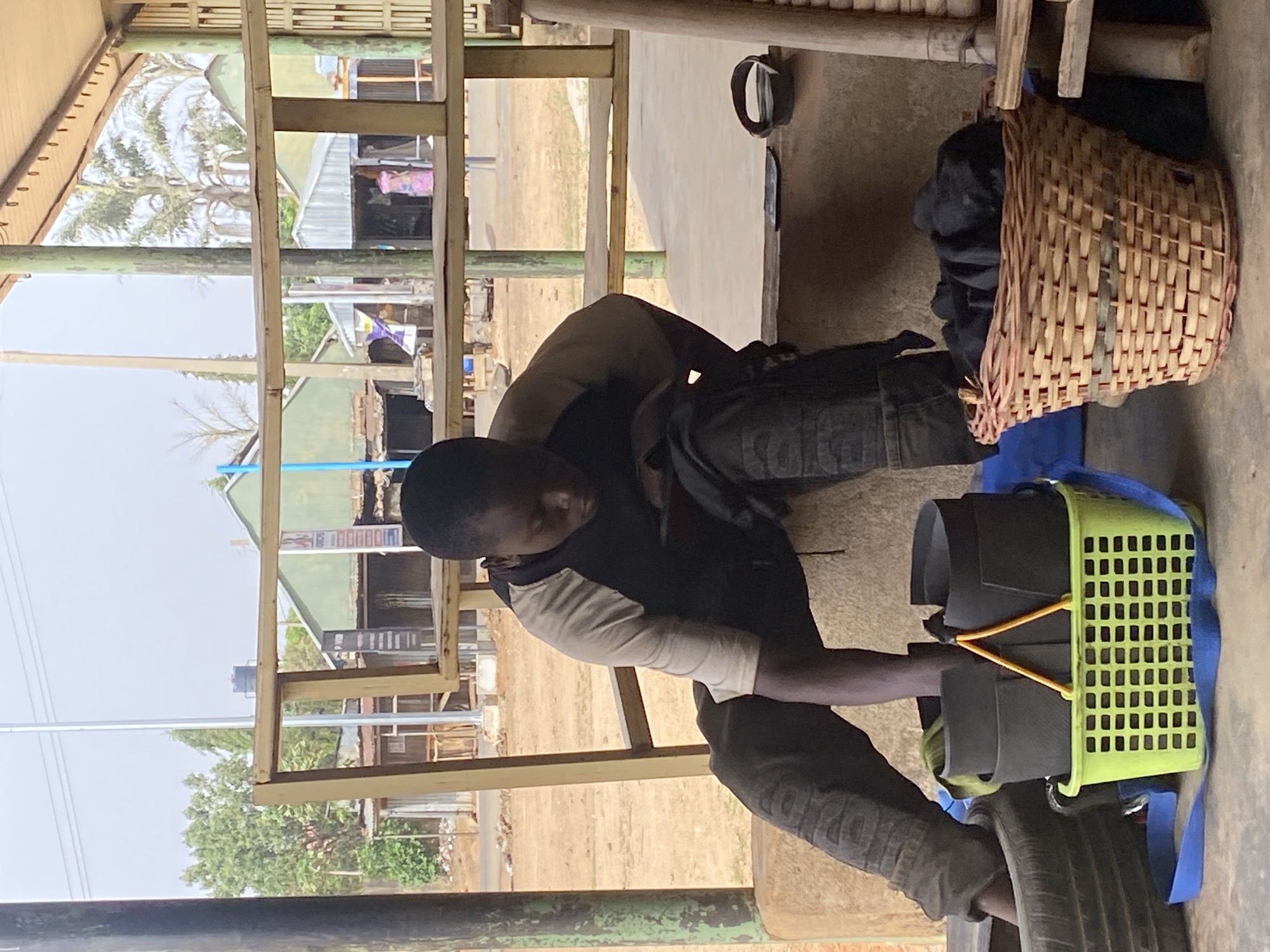 A man sits on a low platform or tire inside what appears to be a partially constructed or open-air structure. He is positioned in front of a green plastic crate that contains some dark material, secured with a yellow bungee cord, and resting on a blue cloth. He is looking at or working with something in his hands near the crate. Beside him is a woven basket with black material inside. The background shows a dirt or sand area with various structures, including buildings and trees. A sign in the distance is visible between the open structure in the foreground. Given the prompt's information, the scene is likely taking place in the city of Jos, Nigeria.