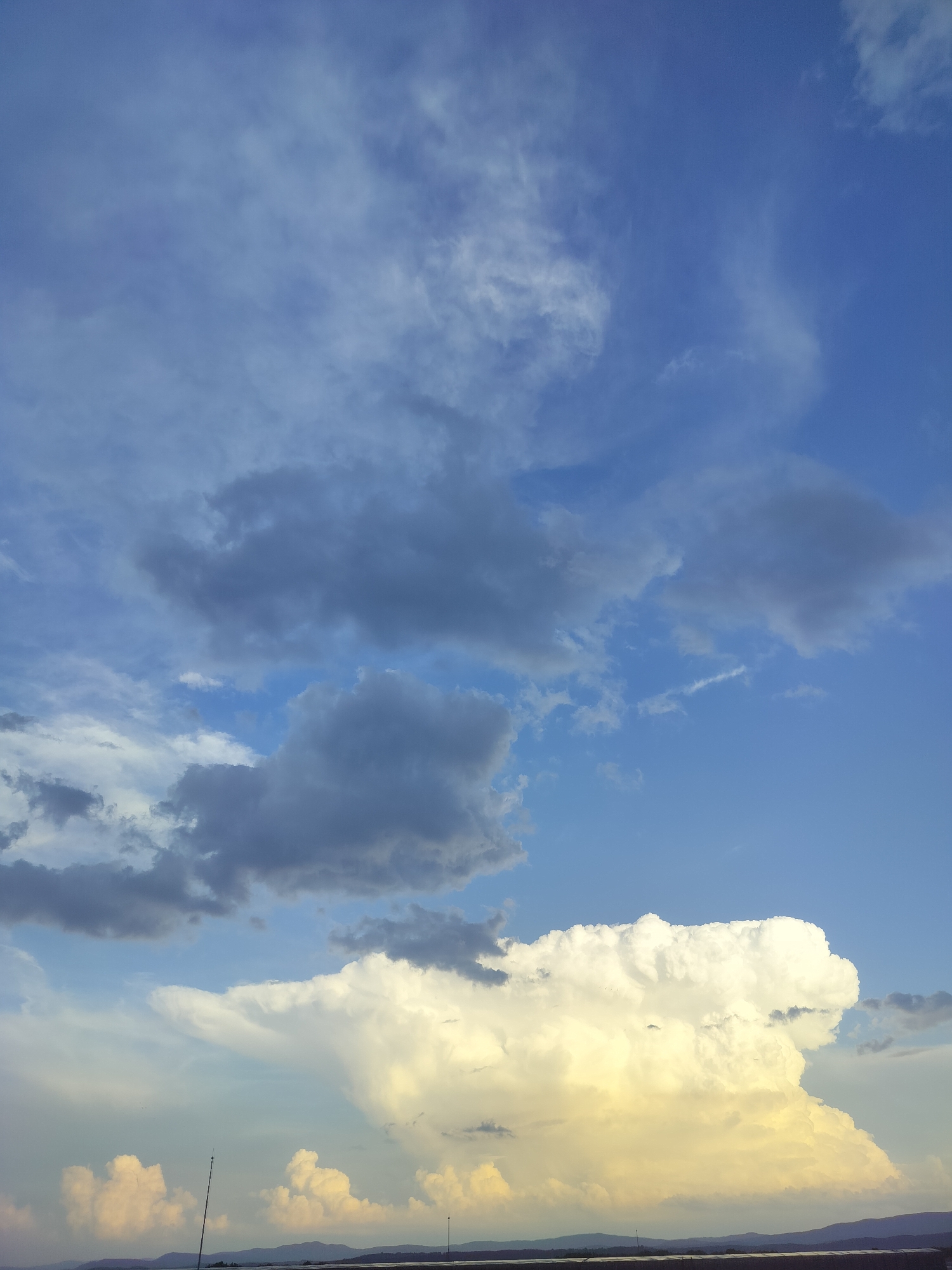 The image displays a vast and dramatic sky over a distant landscape, likely in Gorno Sahrane, Bulgaria. The upper half of the frame shows a gradient of blue sky, from a deeper hue in the upper right to a lighter tone towards the left, dotted with wispy white and scattered grey clouds.
The most striking feature is a massive, towering cloud formation dominating the lower-center of the image. This cloud, resembling a cumulonimbus, exhibits brilliant white tops and a distinct golden-yellow illumination along its lower and central body, suggesting it's catching the warm light of a setting or rising sun. Below it, towards the lower left, are smaller, fluffy white clouds also glowing with the same golden light. Darker, grey clouds are interspersed across the mid-ground sky, contrasting with the brighter blue and illuminated formations.
Along the horizon, a faint line of dark, distant mountains or hills stretches across the width of the image. The very bottom edge shows a dark, relatively flat strip of land or infrastructure, with two thin, vertical poles extending upwards from it. The overall impression is one of a dynamic cloudscape, with the golden-lit cloud being the focal point, suggesting a beautiful late afternoon or early evening. There is no visible text in the image.