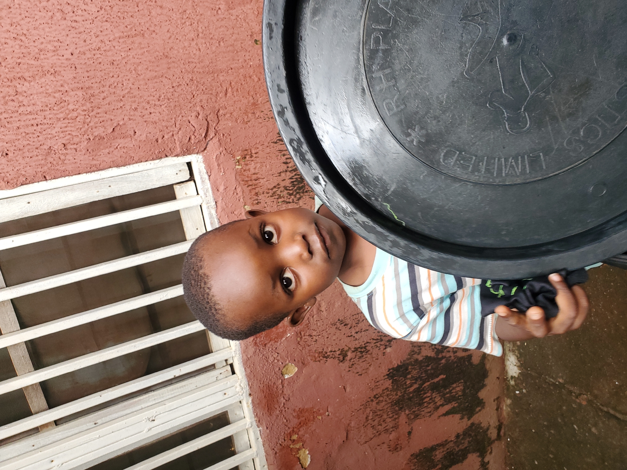 A young boy, estimated to be between 4 and 6 years old, stands outdoors in front of a textured, reddish-brown wall. He is wearing a horizontally striped t-shirt with colors including blue, orange, and white. To his right, a large, dark plastic lid obscures part of the frame. The lid has embossed lettering that appears to read "LIMITED" and "SOLUTIONS" in a circular pattern, and a stylized image of a person running. The boy is looking towards the camera with a neutral expression. Behind him, a window with white bars is visible, suggesting an urban setting. The ground is wet and appears to be paved. The scene is consistent with an outdoor area in the city of Jos, Nigeria.