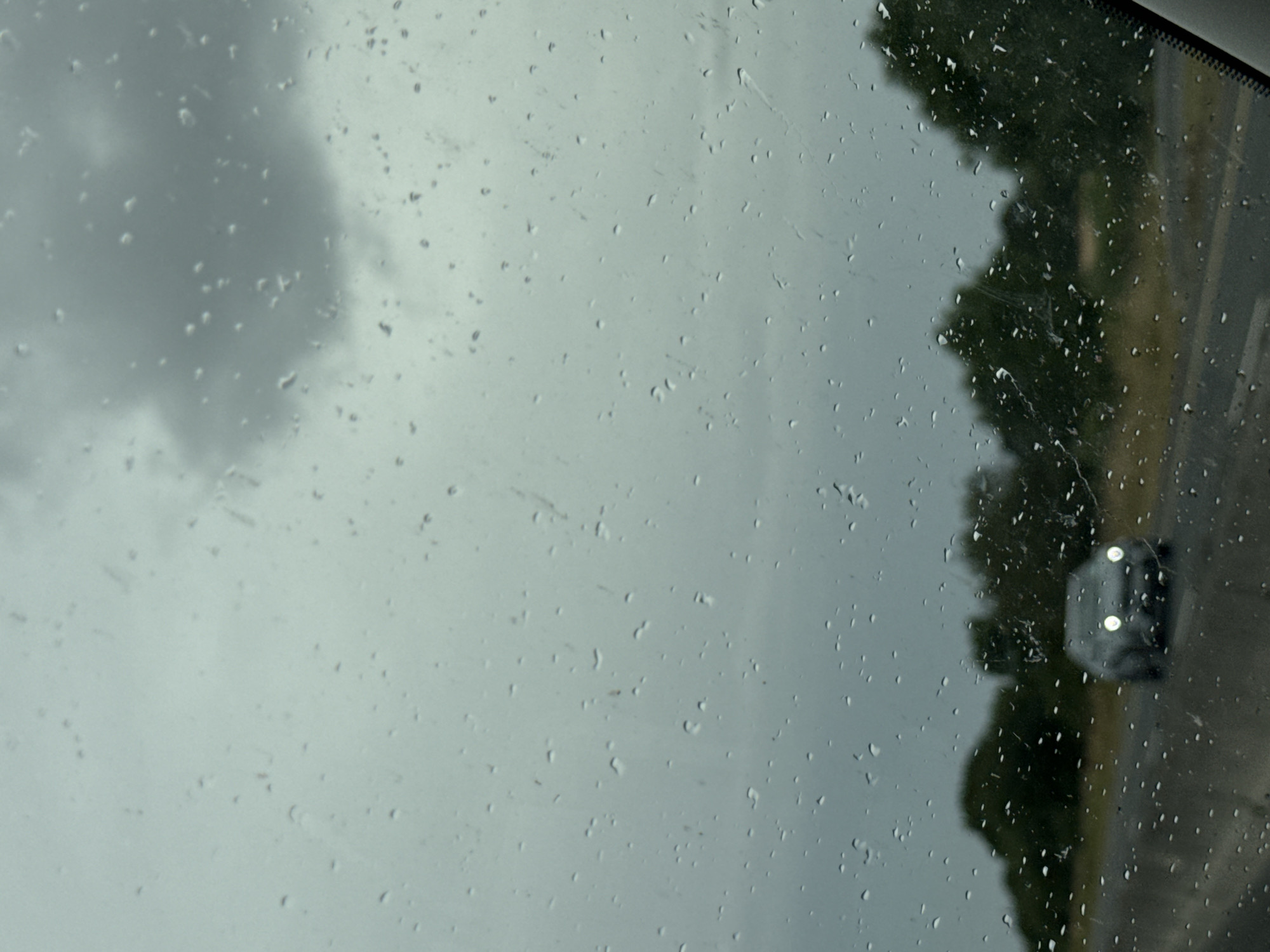 The image depicts a view through a rain-streaked car windshield, looking out at a cloudy sky and a rural landscape. In the foreground, numerous raindrops distort the view. Further out, a dark-colored car with its headlights on is visible on a road, moving towards the viewer. Beyond the road, a line of green trees forms a dense backdrop against the overcast sky. The scene appears to be a roadside view during a rain shower in Craan, Ireland.