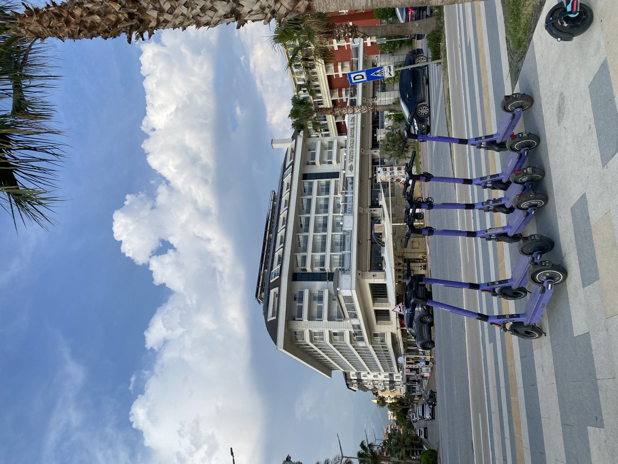 The image, taken in Alanya, Turkey, depicts a vibrant daytime street scene. The perspective is from ground level, looking slightly upwards and across a street.
In the foreground, a row of approximately five purple electric scooters, branded "Beam," are neatly lined up on a grey concrete sidewalk. Some scooters have visible license plate fragments, such as "AL 3327" and "AL 1237." Beside the sidewalk, there's a paved street with a pedestrian crossing marked by alternating grey and yellow stripes.
Across the street, a number of cars are parked, including a dark-colored sedan. A blue road sign with a white 'D' symbol and another symbol indicating a car and pedestrian is visible on the street side. The dominant structure in the midground is a large, modern multi-story hotel building, primarily white with numerous windows and balconies. A clear sign on its facade reads "WHITE GOLD HOTEL & SPA." Behind this main building, other multi-story residential or commercial buildings, some with reddish-brown facades, can be seen, interspersed with palm trees.
The sky, filling the upper portion of the frame, is a clear blue with large, voluminous white cumulus clouds concentrated on the right side and thinner, wispy clouds on the left. The fronds and trunks of palm trees partially frame the view from the top and left edges, suggesting a tropical or Mediterranean climate. The lighting indicates it is a sunny day. No people are prominently visible in the scene, which appears to capture a moment of urban tranquility.