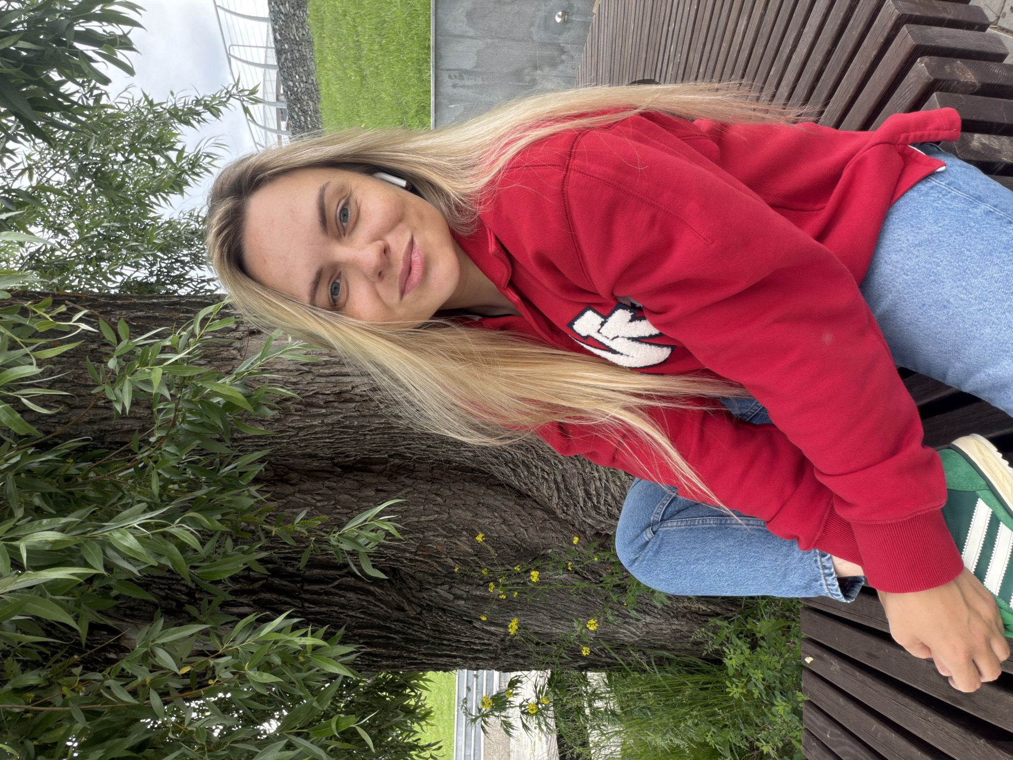 A young woman with long blonde hair and blue eyes is sitting on a wooden bench in a park. She is wearing a red hoodie and blue jeans, and her green sneakers are visible. The woman is smiling and looking at the camera. She has a white wireless earbud in her right ear. She is sitting next to a large tree with rough bark and green leaves. Behind her, there is a grassy slope and a stone wall. The sky is overcast. The scene is located in Votkinsk, Russia.
