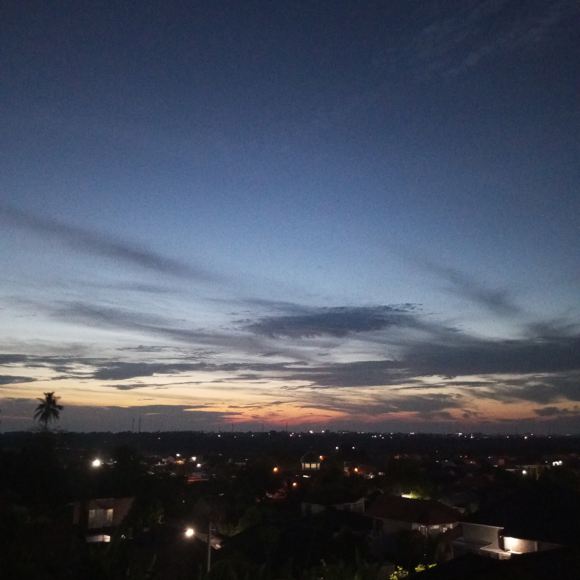 The image depicts a cityscape at dusk, with a view over what appears to be a residential area. The sky is a gradient of blues, greys, and oranges at the horizon, indicative of a sunset or sunrise. Clouds are streaked across the sky. Below the horizon is a darkened landscape dotted with sporadic lights, suggesting street lamps or lights from buildings. The landscape is flat with some vegetation, including what appears to be a palm tree on the left of the image. The low density of lights suggests a suburban or semi-rural setting. Without specific landmarks or geographic features visible, it is impossible to confirm the exact location in Lhokseumawe. No people are visible.