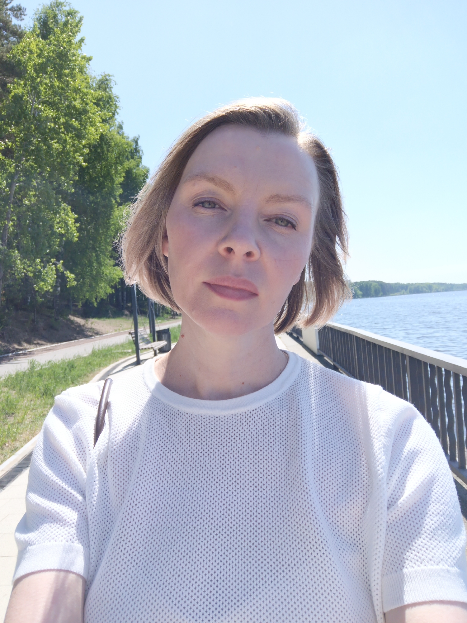 A fair-skinned woman with short, light-blonde hair stands facing the camera, her gaze directed towards the viewer. She wears a white, mesh-like T-shirt. Her arms are crossed in front of her. Behind her is a paved walkway bordered by lush green trees on one side and a metal railing overlooking a body of water. The water stretches out to the horizon under a clear, light blue sky. The location is likely in the city of Chaikovsky, Russia, based on inference.
