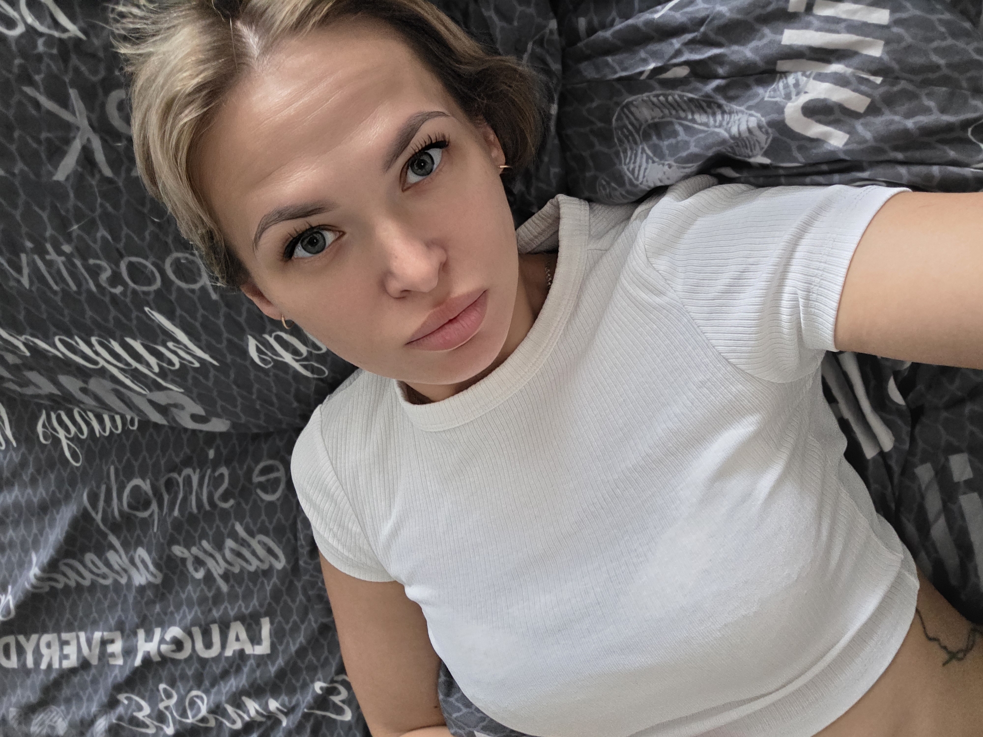 A woman with short blonde hair and blue eyes is lying down on a grey bedspread with white writing on it. She is wearing a white, short-sleeved t-shirt. The image is a close-up selfie, focusing on her face and upper body.
The location cannot be determined from the image alone. The mention of Chaikovsky, Russia, is likely extraneous.