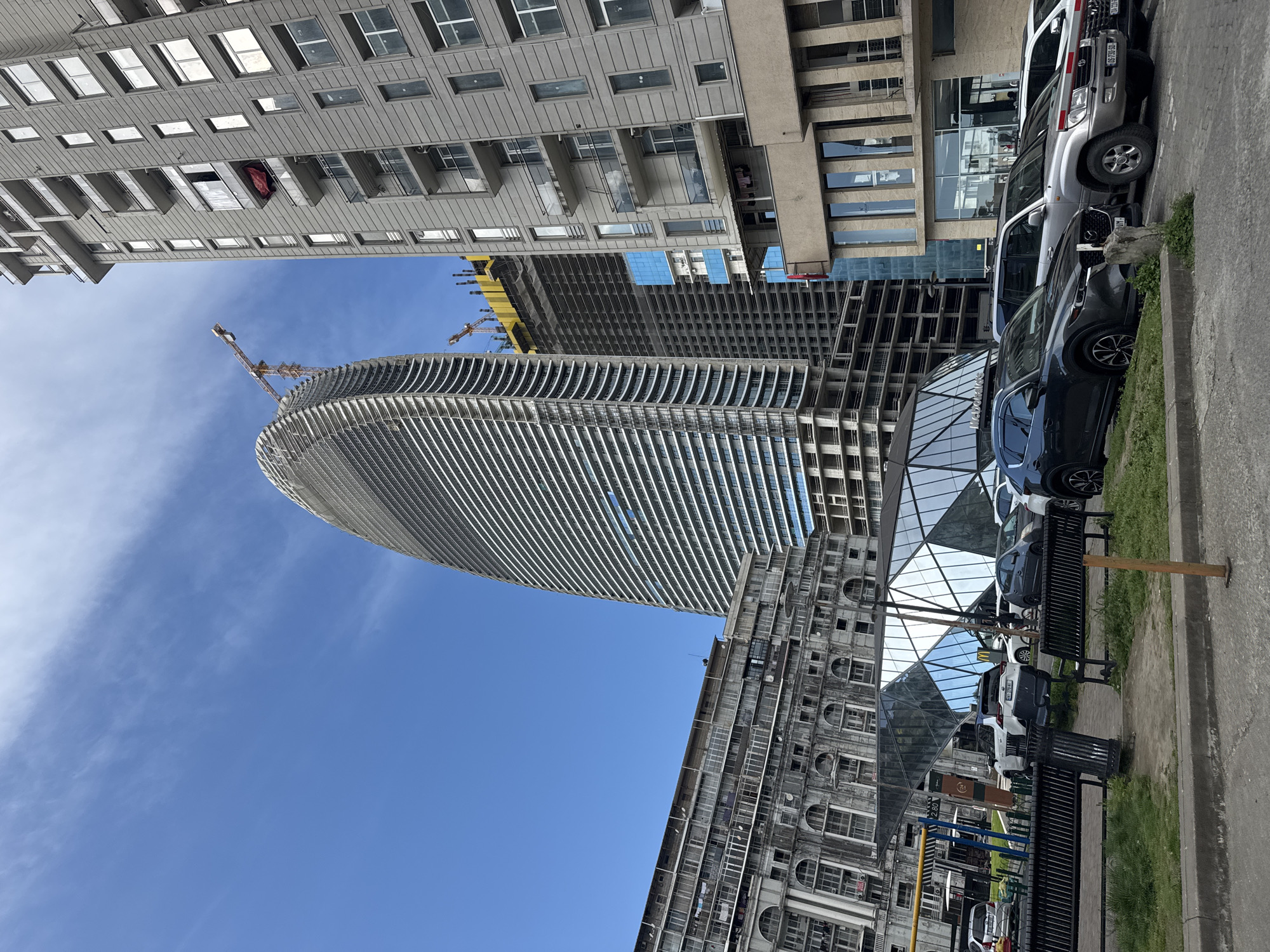 The image captures an urban street scene under a bright, clear blue sky with wispy white clouds. The primary subject is a tall, slender skyscraper under construction, featuring a distinct curved facade with numerous horizontal fins or balconies. A large yellow construction crane is visible near its unfinished upper sections.
To the right of the skyscraper, an older, multi-story building stands with a more ornate, classical design, characterized by rows of arched windows and balconies. At its base, a modern, faceted glass structure reflects the sky and surrounding buildings. A partial "McDonald's" logo is visible on a sign within this glass structure. To the left, a modern, flat-faced building with rectangular windows and balconies suggests residential or office use. Further back, another modern building with reflective glass facades is visible.
Along the curb in the foreground, several vehicles are parked, including a silver Toyota Land Cruiser SUV (with a partial license plate "099 T-R-080") and a dark Mazda CX-5 SUV (with a partial license plate "UU-U 164"). Black metal benches and a trash can are present on the paved sidewalk, next to a small strip of grass. A yellow sign with "250" in black text is visible on the older building.
The scene depicts ongoing urban development, contrasting modern construction with older architecture, suggesting a bustling city environment during bright daylight.