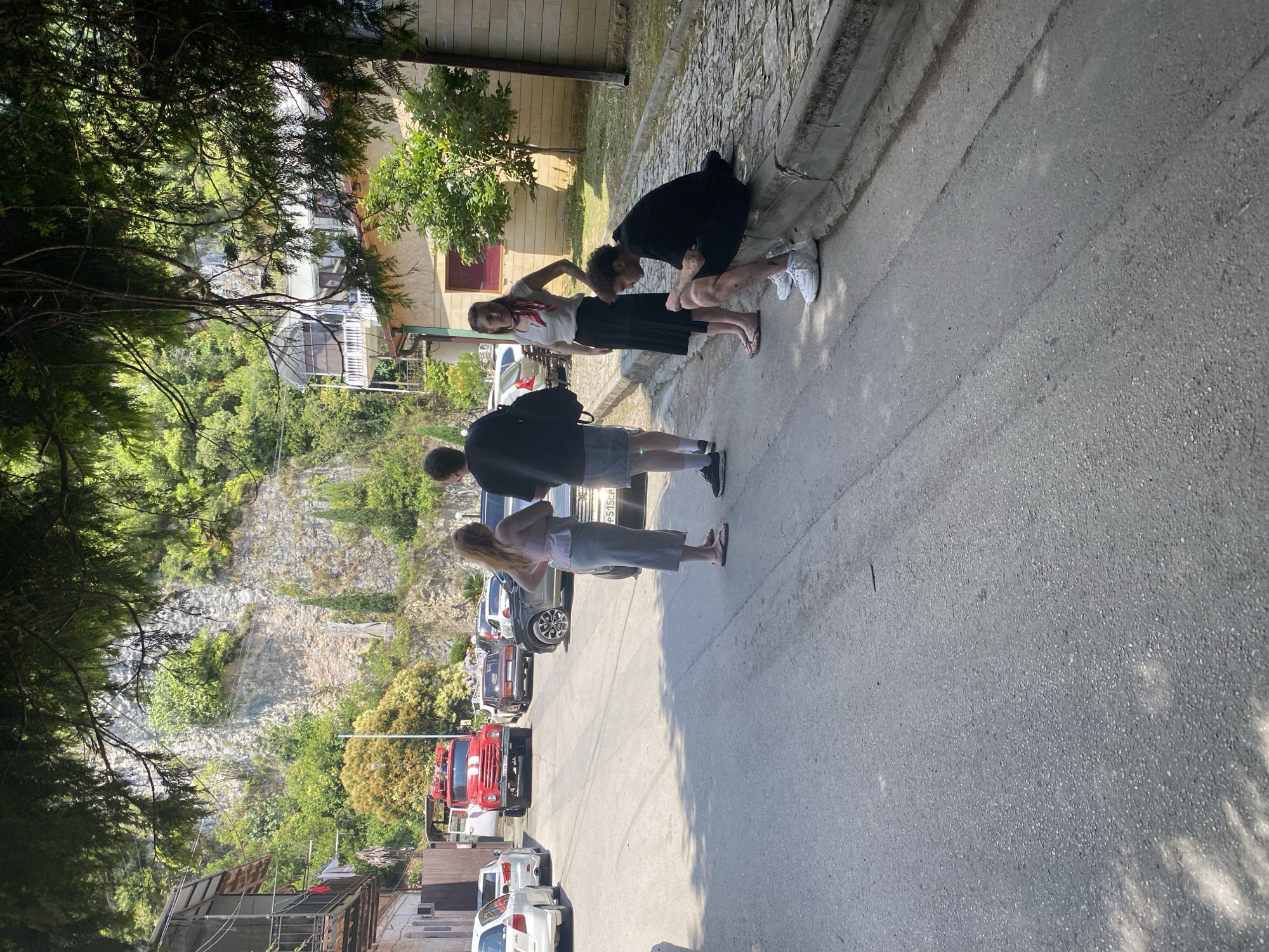 In a sunlit, outdoor scene, likely a street in an urban area, four individuals are gathered. A red fire truck is parked on the asphalt road, and several cars are visible in the background. The environment features a steep, rocky incline covered with greenery, suggesting a mountainous or hilly terrain. Buildings with balconies and balconies can be seen along the left side of the road, and a stone-paved walkway runs parallel to the road on the right.
The individuals are positioned in different areas of the scene. Two people, a woman and a man, are standing and facing each other near the center of the road. The woman is wearing a pink top and a long gray skirt, with her hands on her hips. The man, in a black t-shirt and shorts, stands with his back to the camera. Further to the right, a woman in a white top and black skirt is standing and looking at a young man who is sitting on the curb, wearing a black t-shirt and shorts. The young man is looking down, and his legs are dirty. The other individuals appear to be casually dressed. The overall atmosphere is one of a brief pause or gathering on a street with a backdrop of natural and built environments.